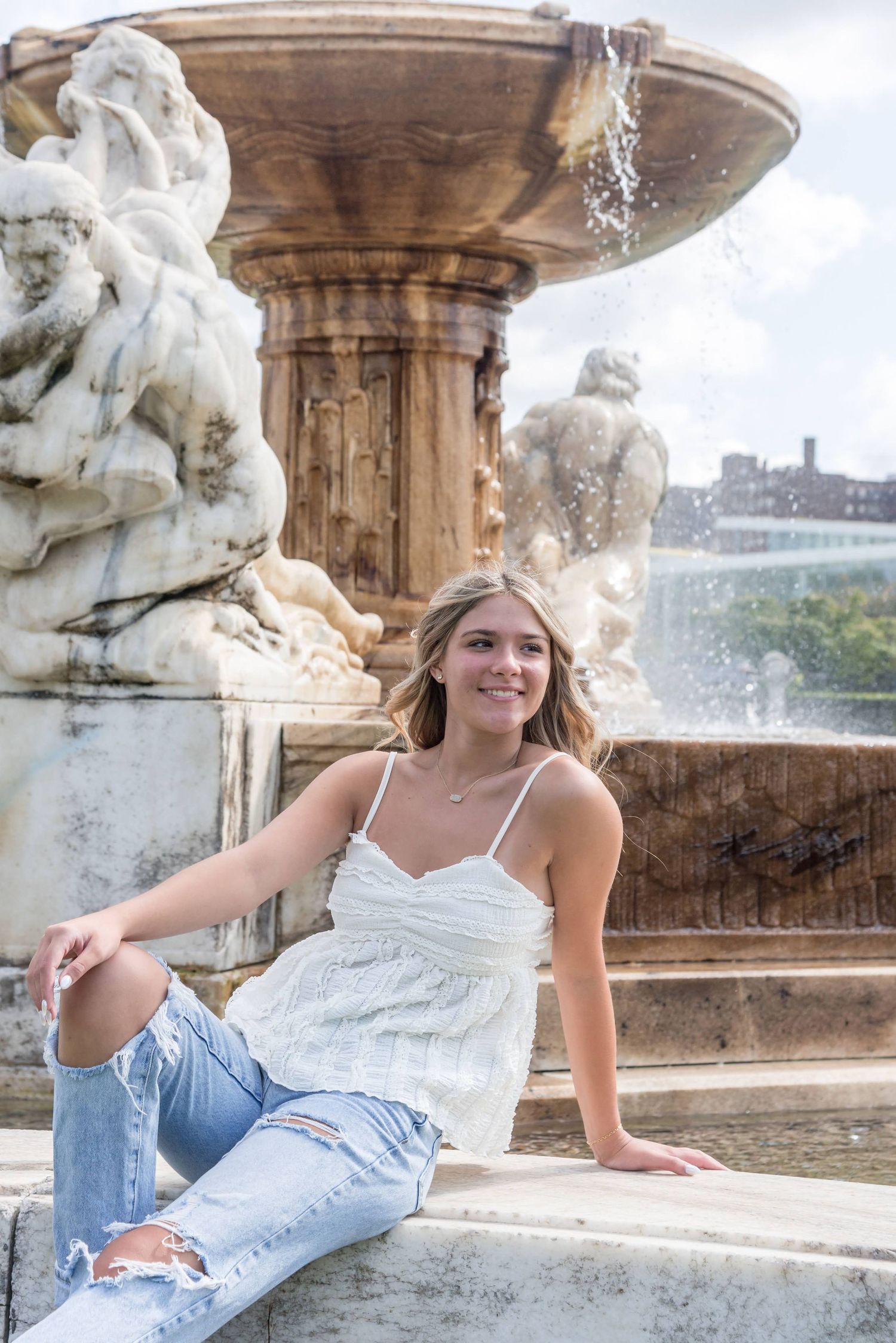 Casual summer style with white top and ripped jeans posed by decorative stone fountain.