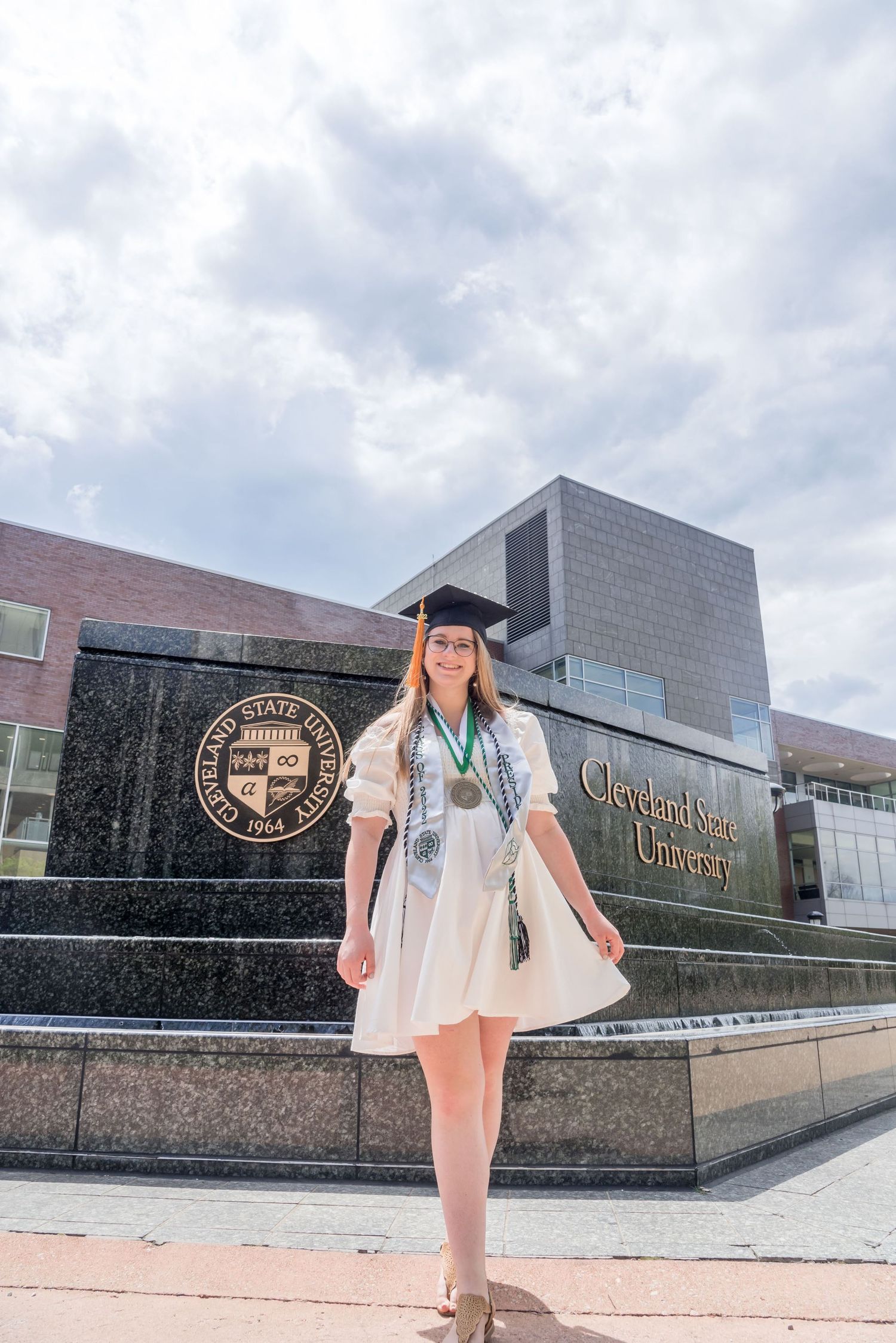 Student stands in front of modern university building with graduation gown.