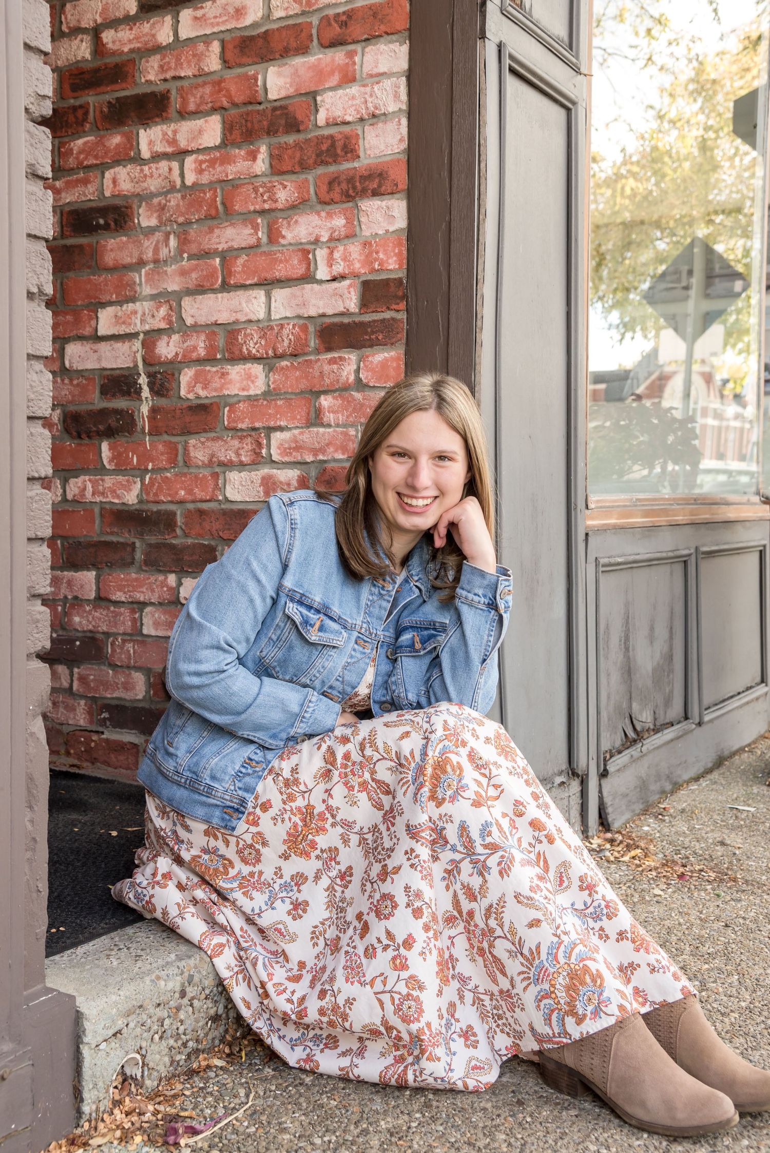 A person in denim jacket and floral dress sits casually against a brick wall.