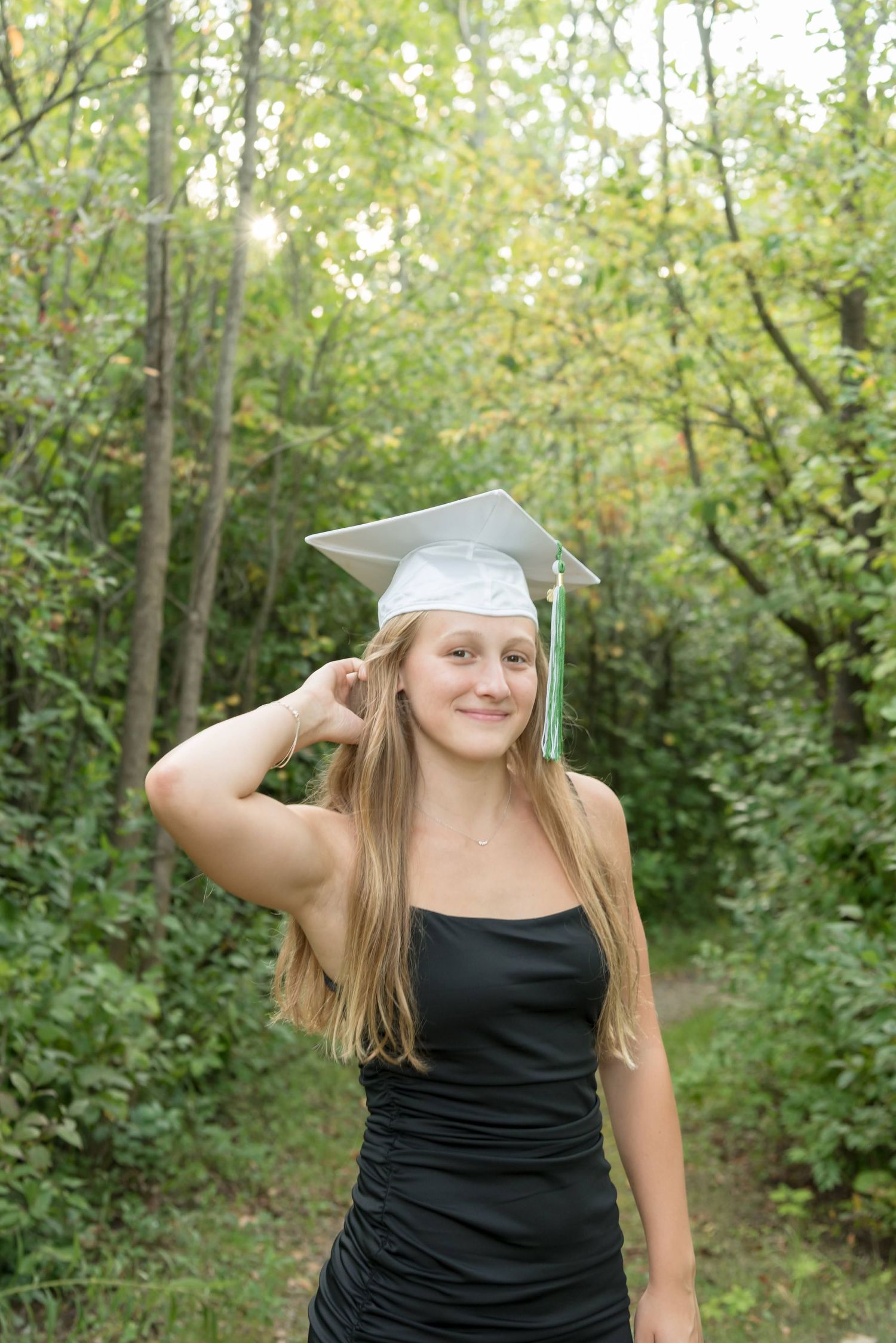 Graduate in black dress and white cap posing playfully in wooded outdoor setting.