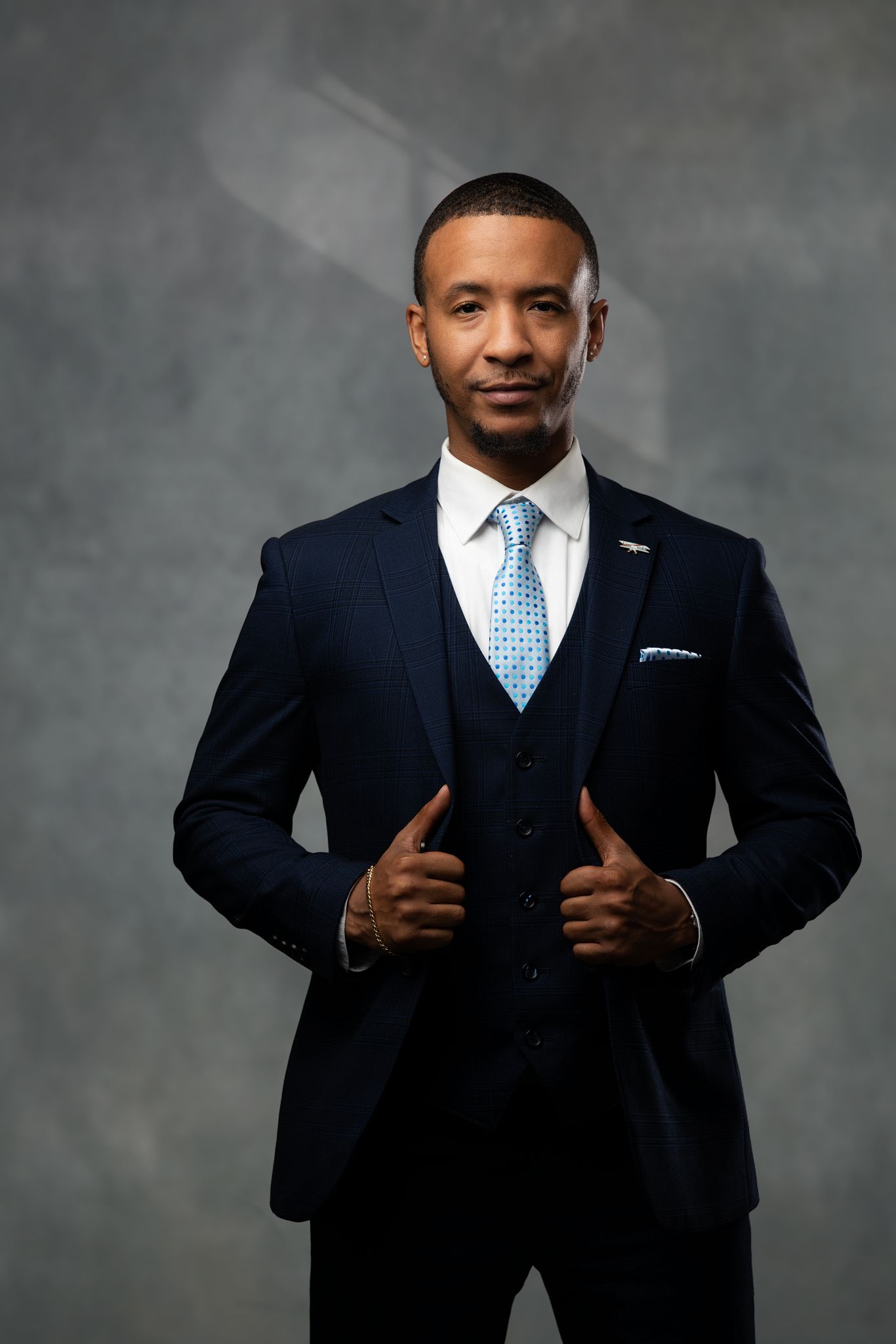 Professional wearing a navy three-piece suit and light blue patterned tie poses against a grey backdrop.