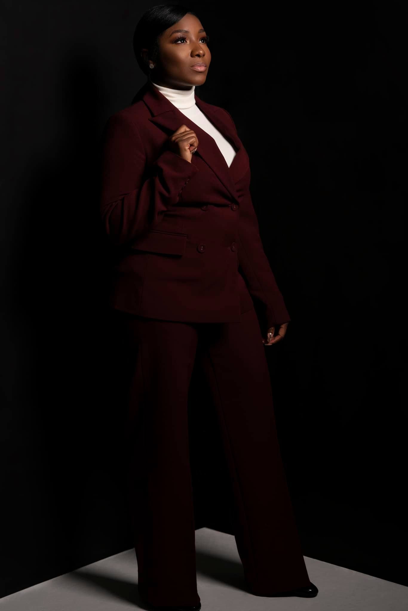Dramatic lighting highlights burgundy pantsuit in dark studio setting with confident pose.