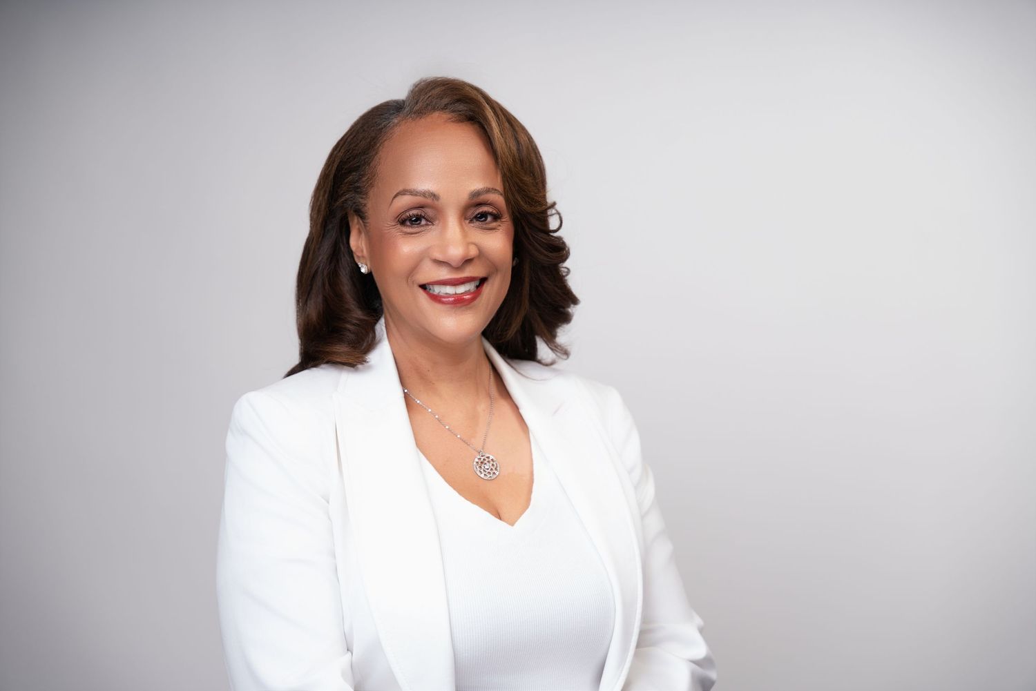 Professional headshot of someone in a white blazer smiling warmly against a light gray background.