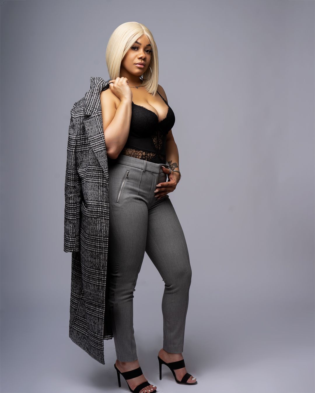 A model poses in a elegant outfit featuring a gray plaid blazer, black top and gray pants with high heels.