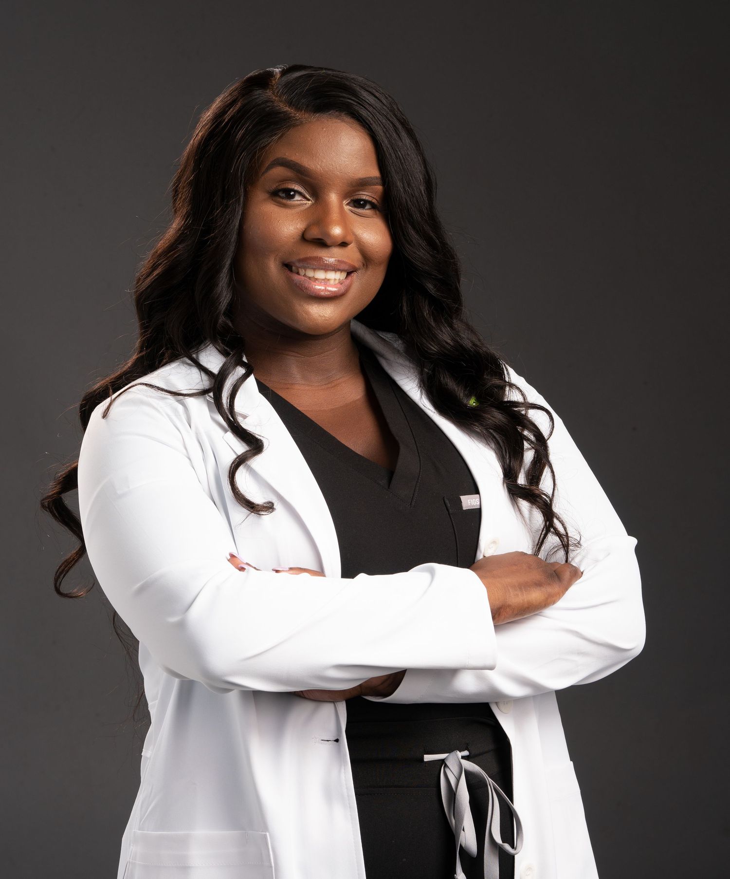Healthcare professional in white coat and black scrubs poses with confident smile.