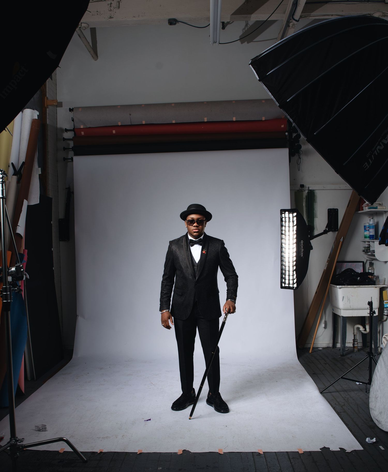Person in classic black tuxedo and hat poses in photo studio with lighting equipment visible.