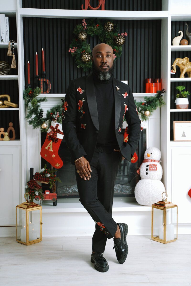 Festive holiday scene with person in black suit posing by decorated fireplace with stockings and wreaths.