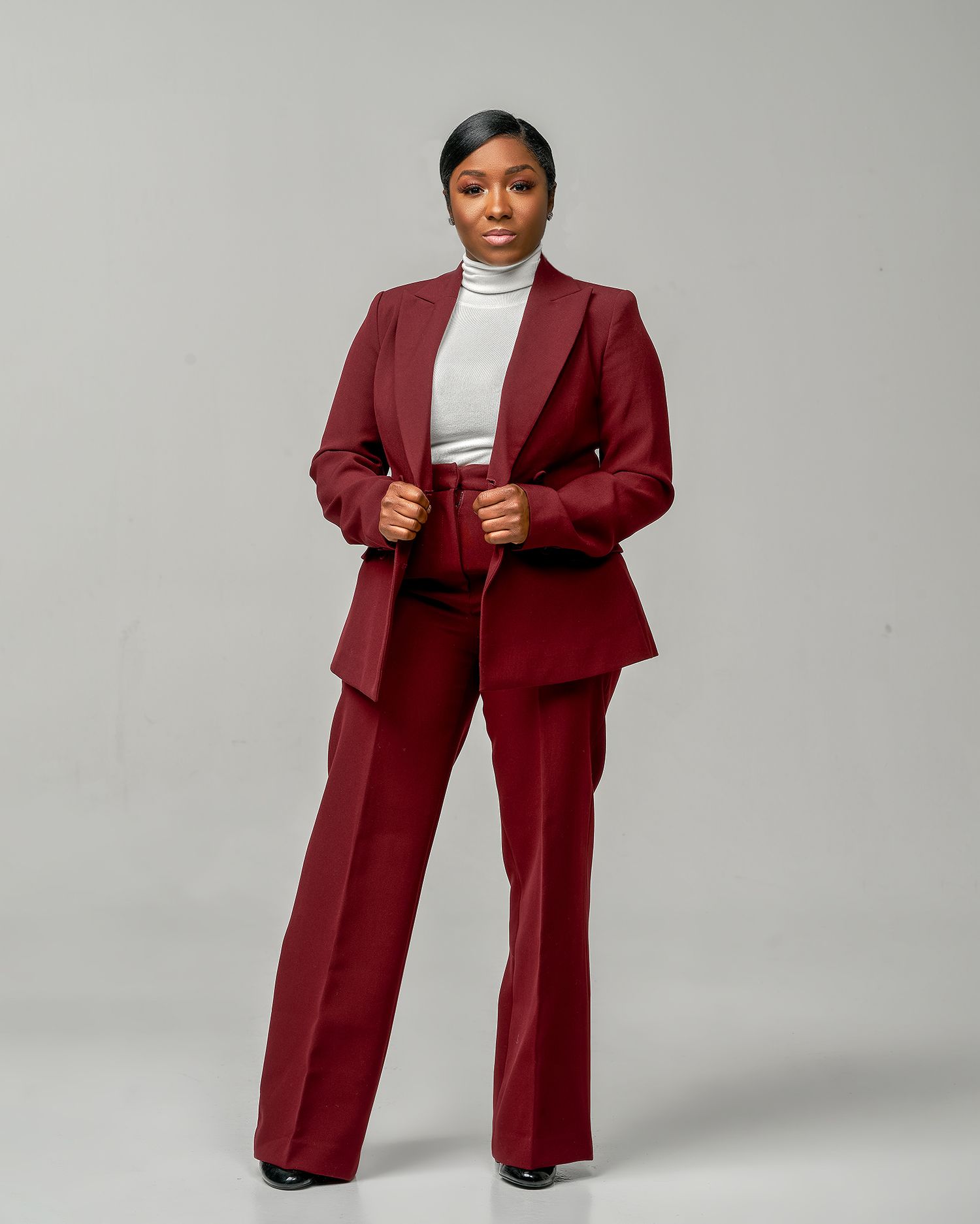 A person wears a burgundy business suit with wide leg pants and white turtleneck in a professional studio setting.