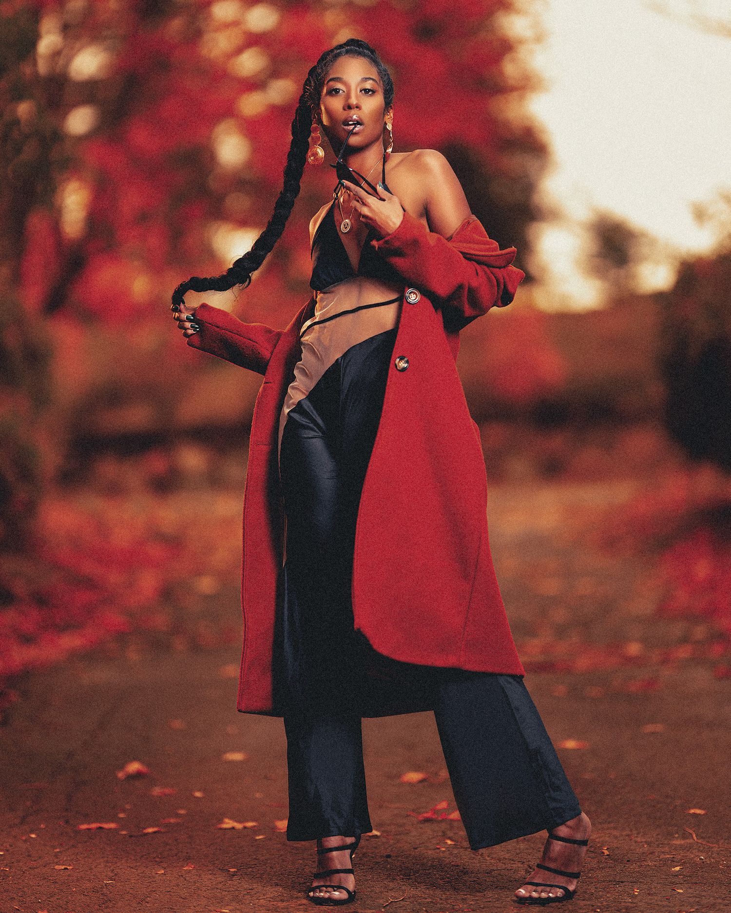 A woman in a red coat and black dress poses gracefully against an autumn backdrop with vibrant red foliage.