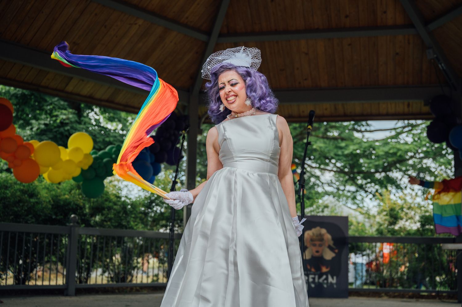 A photo of the drag queen, Mia Moore, dressed in a white dress and waving a flowy rainbow fan.
