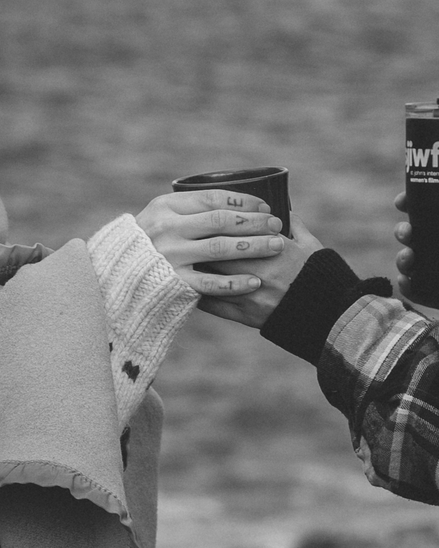 A black and white photo of a hand reaching out and accepting a mug from another hand with knuckle tattoos that say 