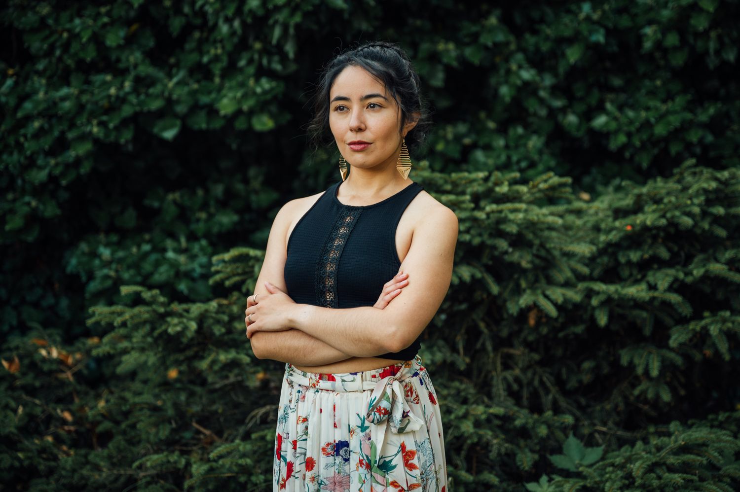 A photo of a human standing strong, with their arms crossed in front of them. They are standing in front of greenery.