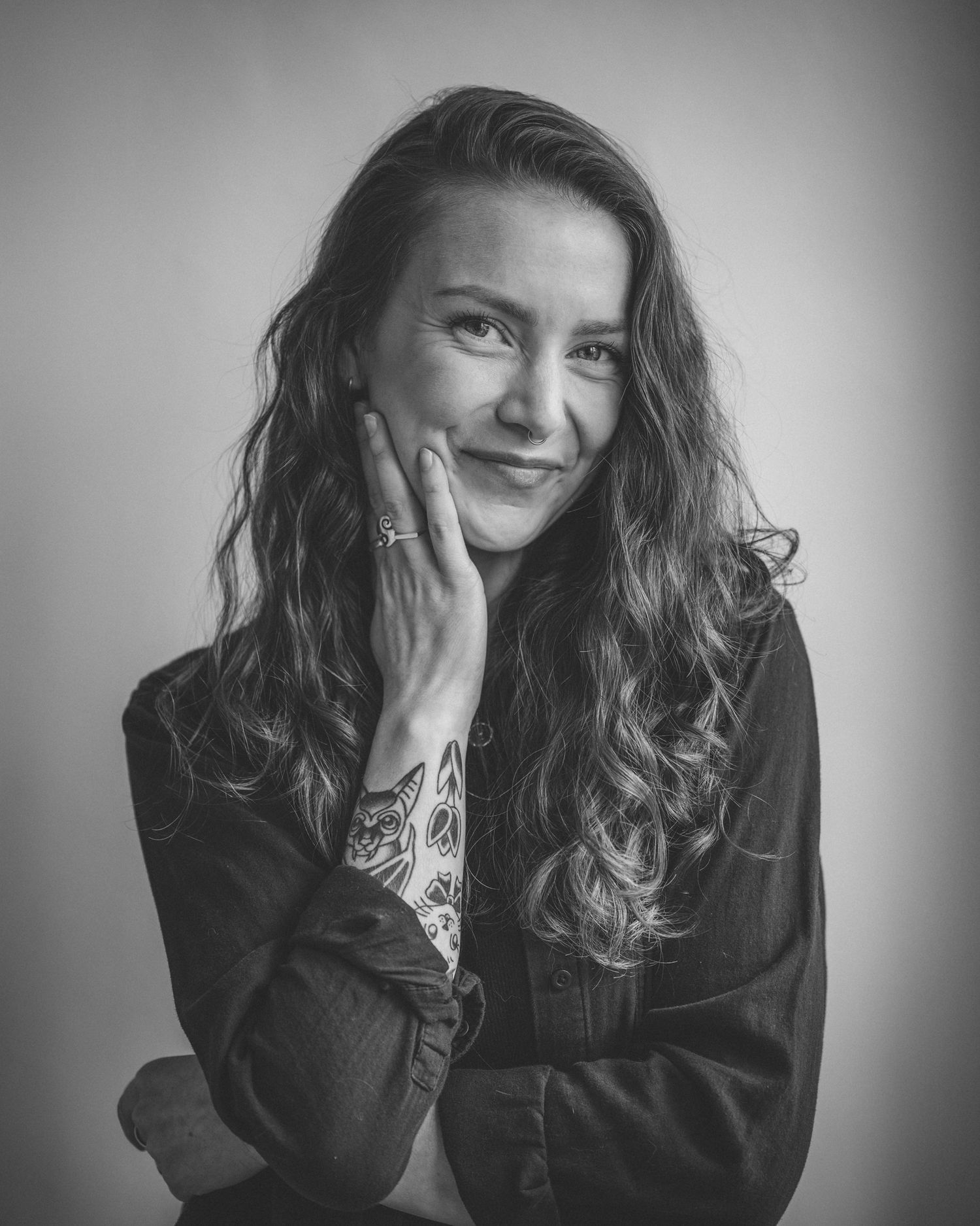 A black and white photo of me smiling at the camera. I have long wavy hair and tattoos. One hand is gently touching my face.