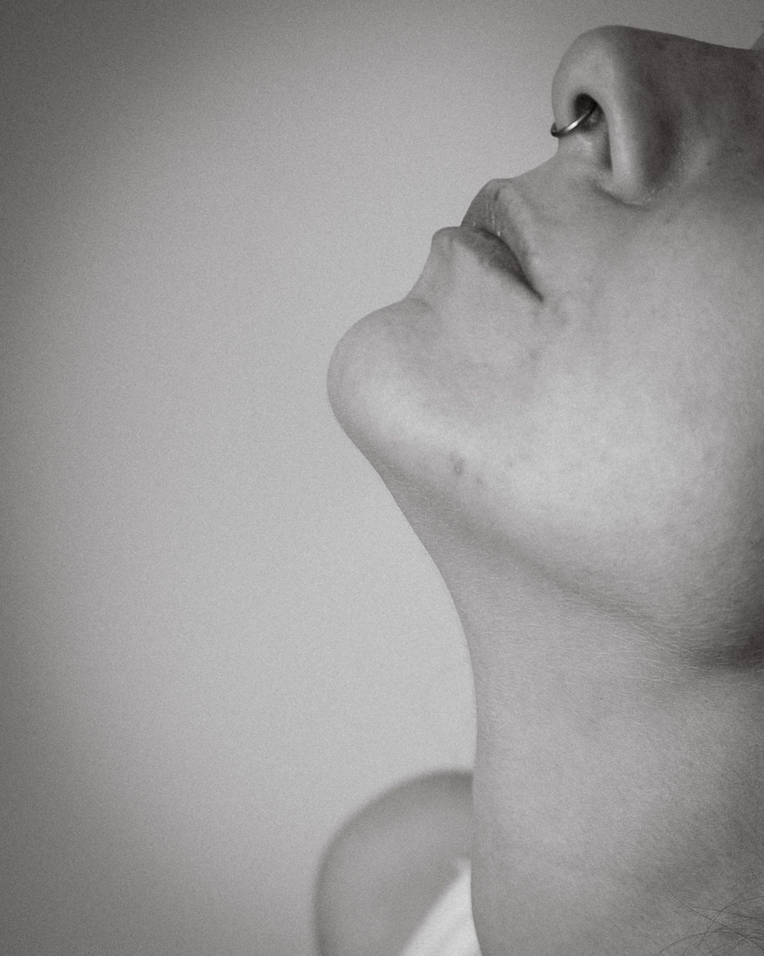 A black and white photo of the bottom of a face, with just the nose (with a septum piercing) and chin visible. The head is tilted back.