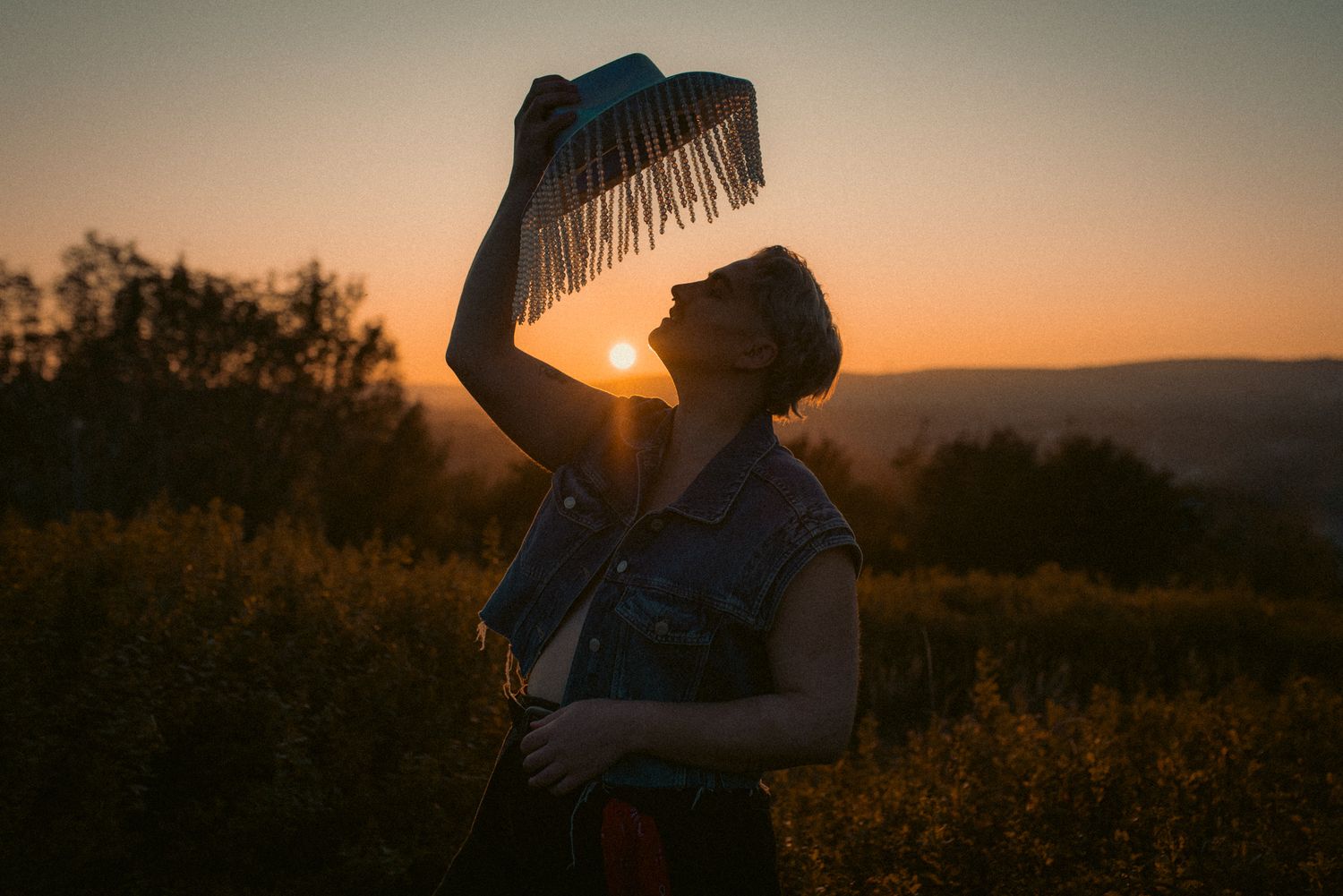 A photo of a human in a denim vest holding up a cowboy hat with strings of beads hanging all around the brim. It's sunset and warm.