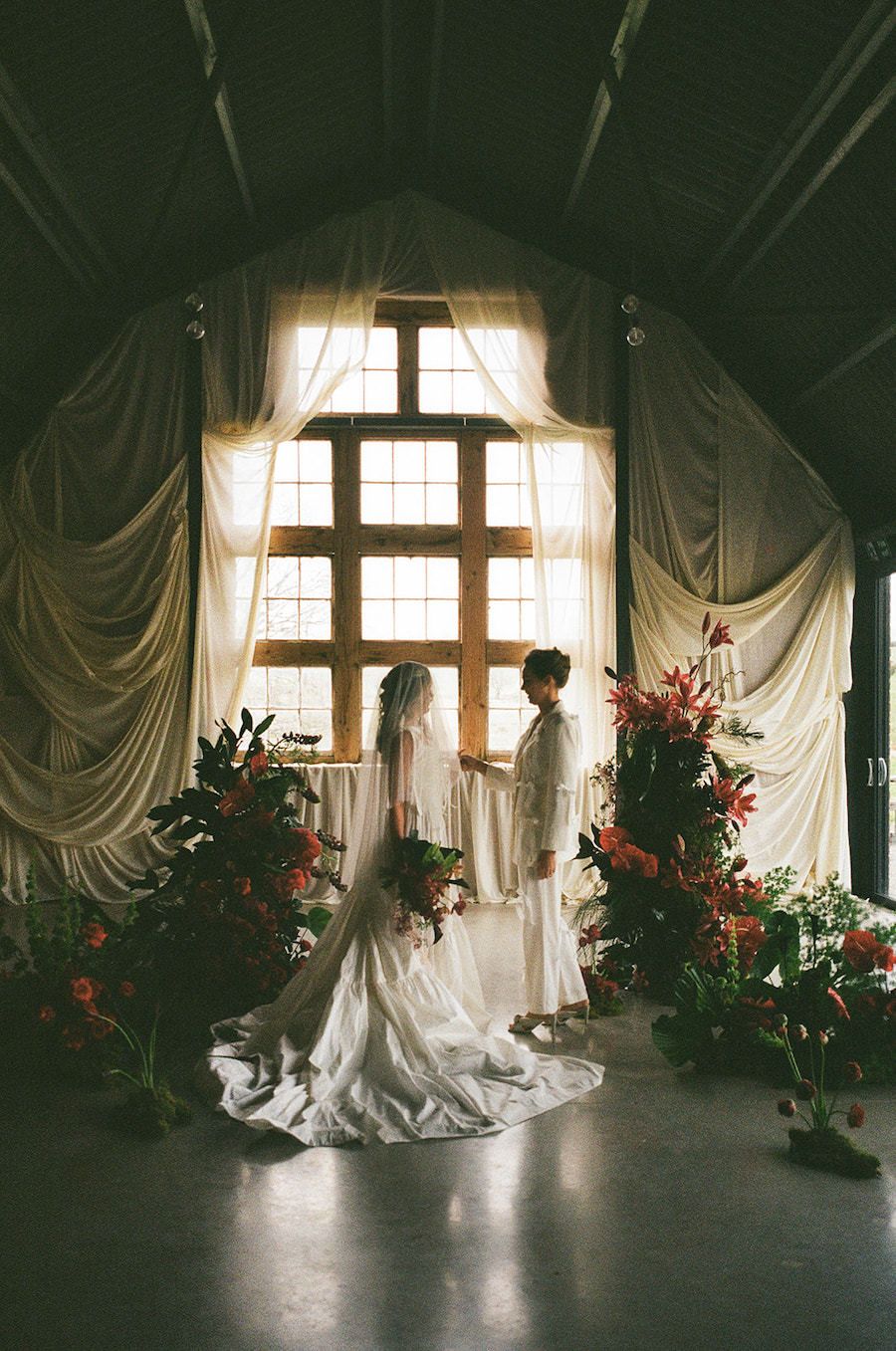 Same sex couple stand at the alter at The Giraffe Shed wedding venue in Newtown, Wales. 35mm Film. Red Flowers decorate the room.