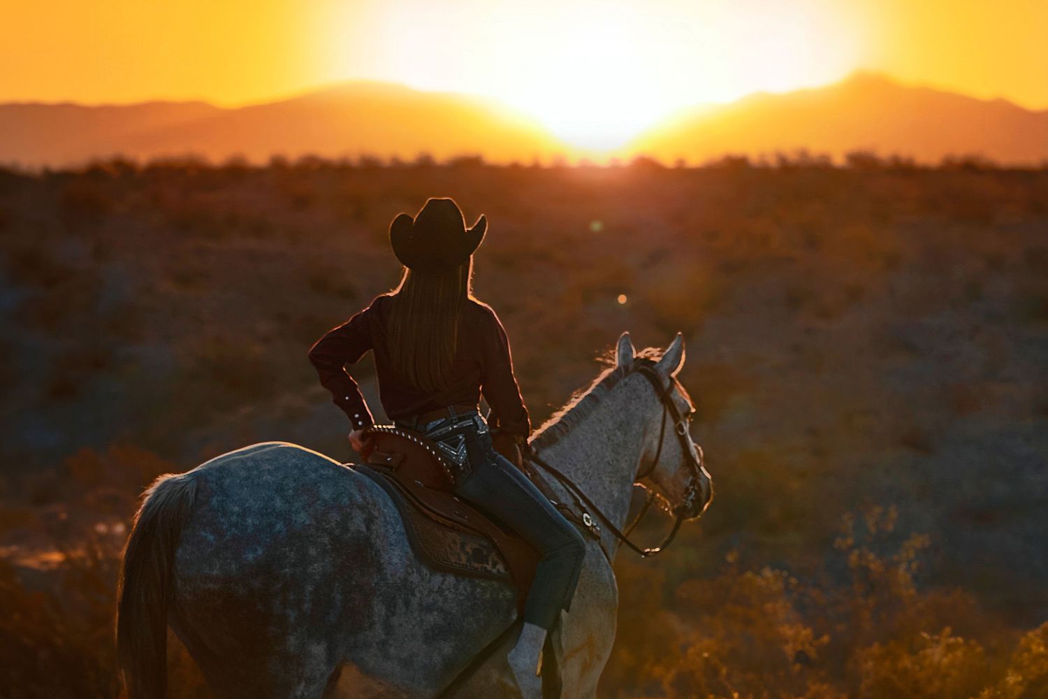 Silhouetted cowgirl on horseback against dramatic orange sunrise in desert landscape. Editorial Portrait, Lifestyle Photography.