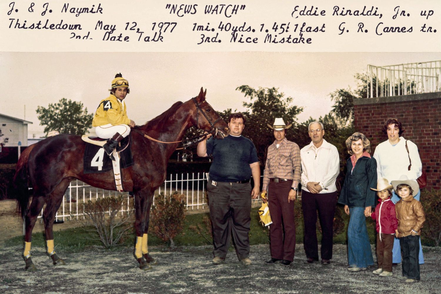 A group of people gathered around a brown horse on a racetrack in a vintage photo from 1977.