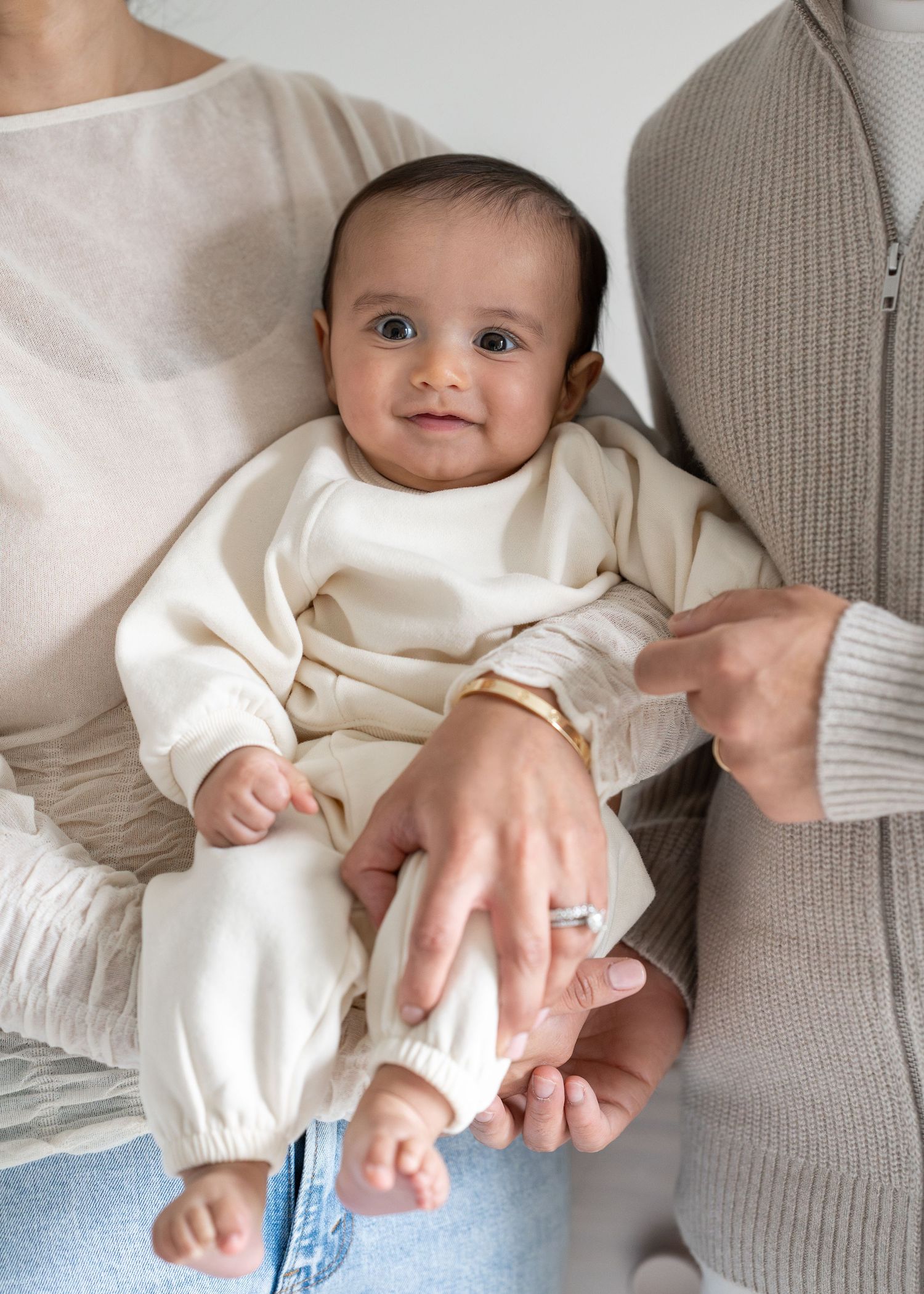 A baby in a cream-colored outfit holds hands with someone wearing a gray sweater while sitting on a white surface.