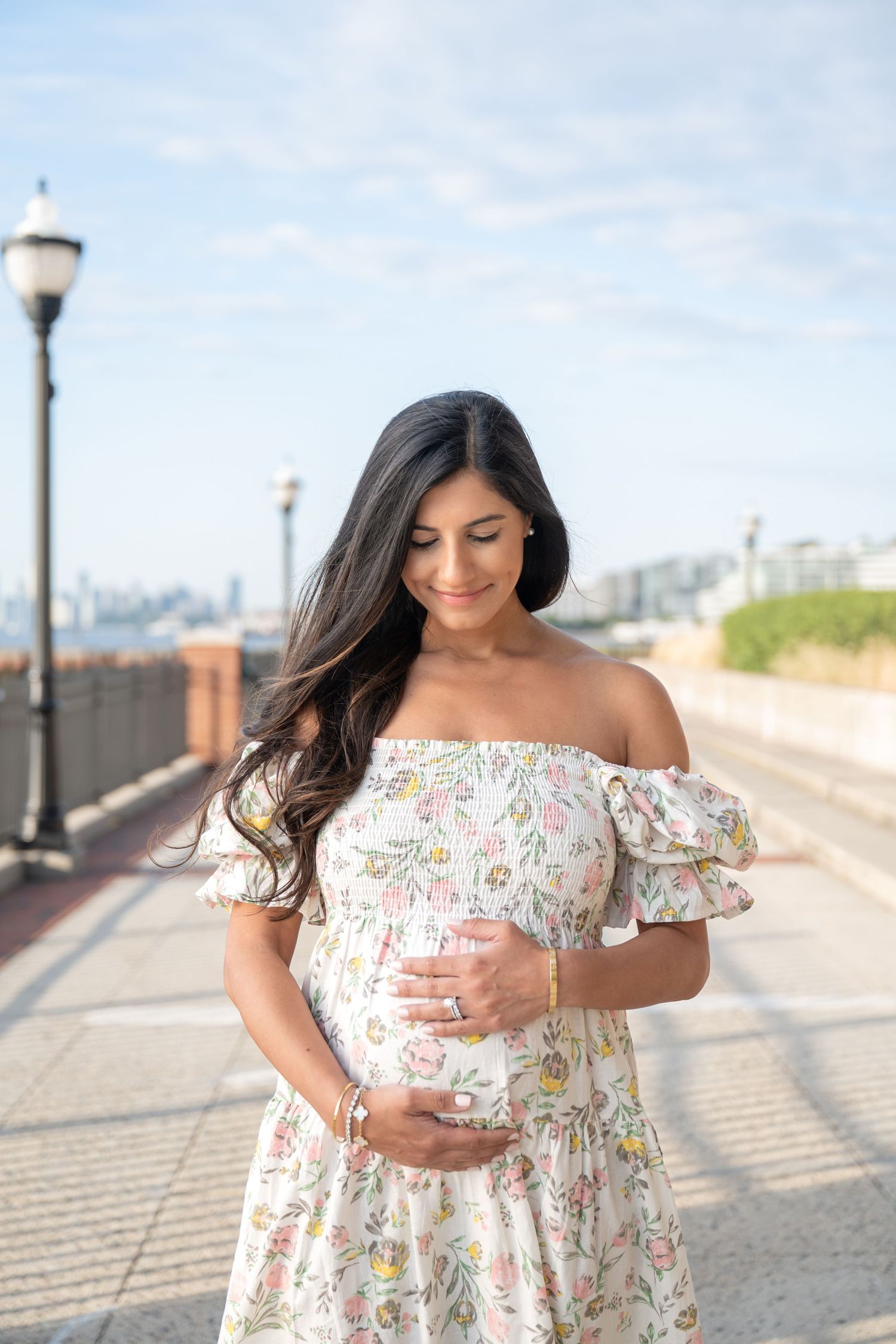 A pregnant woman in a floral off-shoulder dress stands on a sunny walkway with a lamp post and city skyline in background.