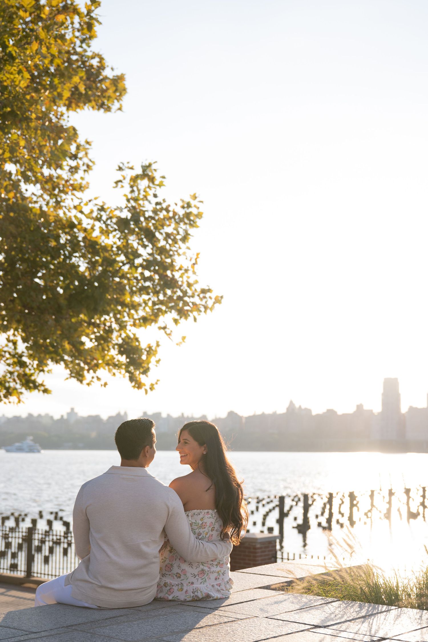 A couple sits together by the waterfront at sunset with golden trees in the background.
