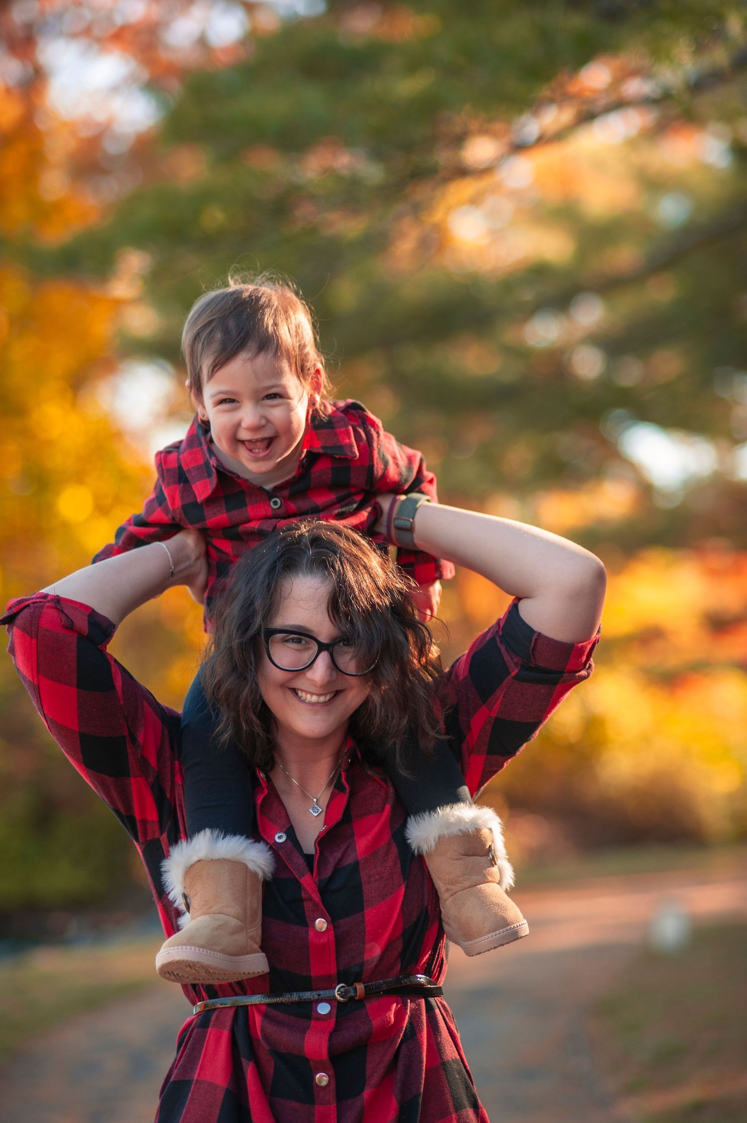 A person in a red and black buffalo plaid shirt poses playfully in autumn sunlight with fall foliage in the background.