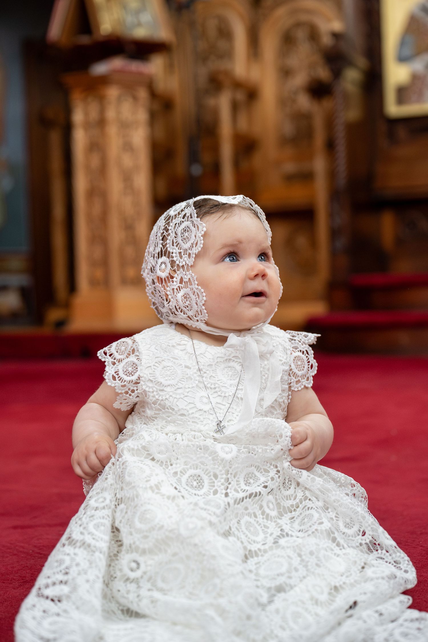 Baby girl in white lace baptism gown and bonnet sitting on red carpet in church.