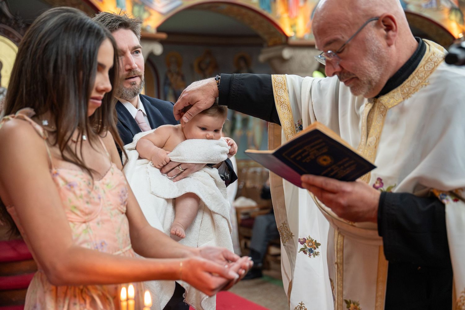 Priest performing a Christian baptism ceremony on an infant held by parents inside a decorated church.
