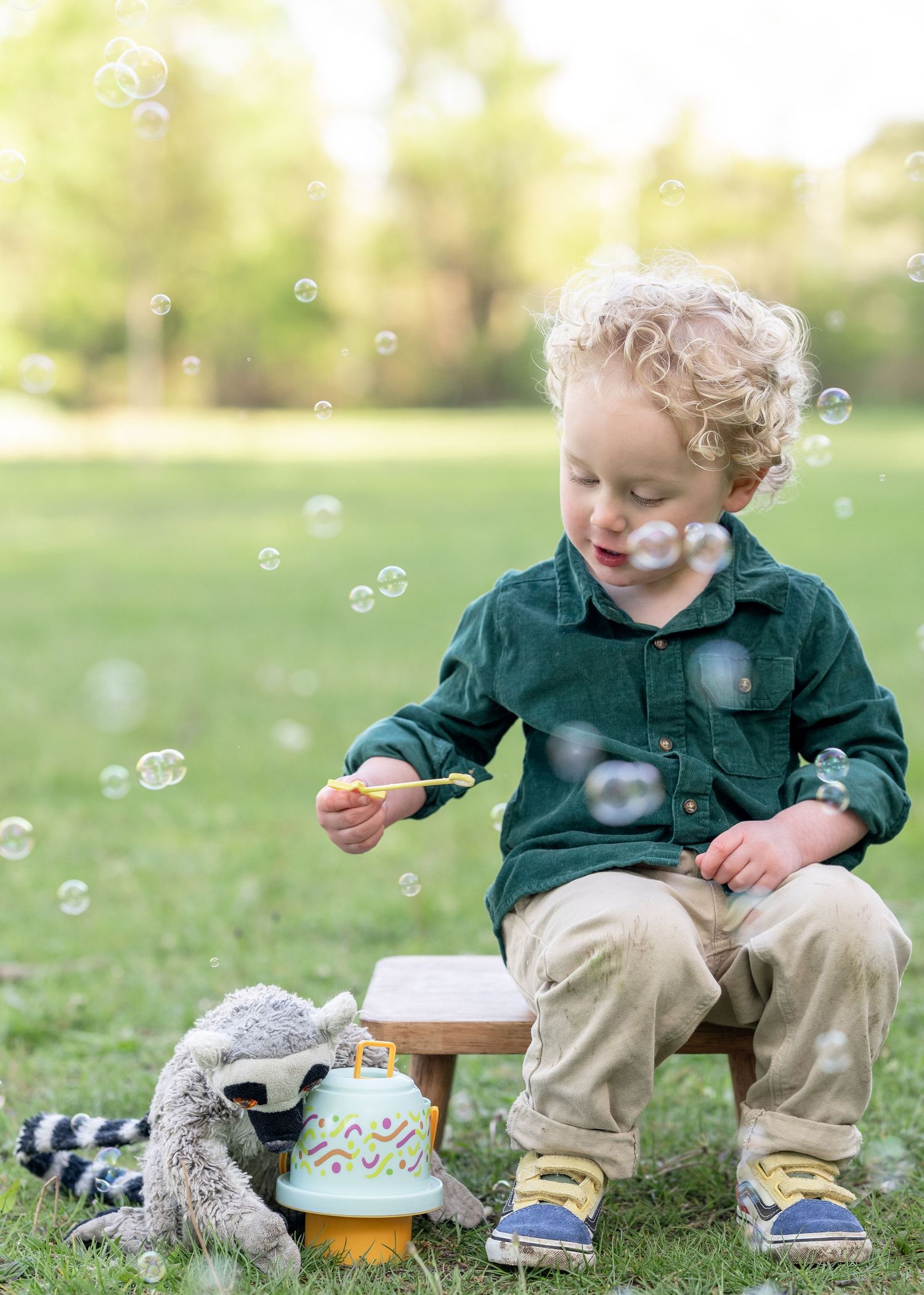 Toddler boy blowing bubbles outdoors, sitting on small stool with toy birthday cake and stuffed raccoon on grass.
