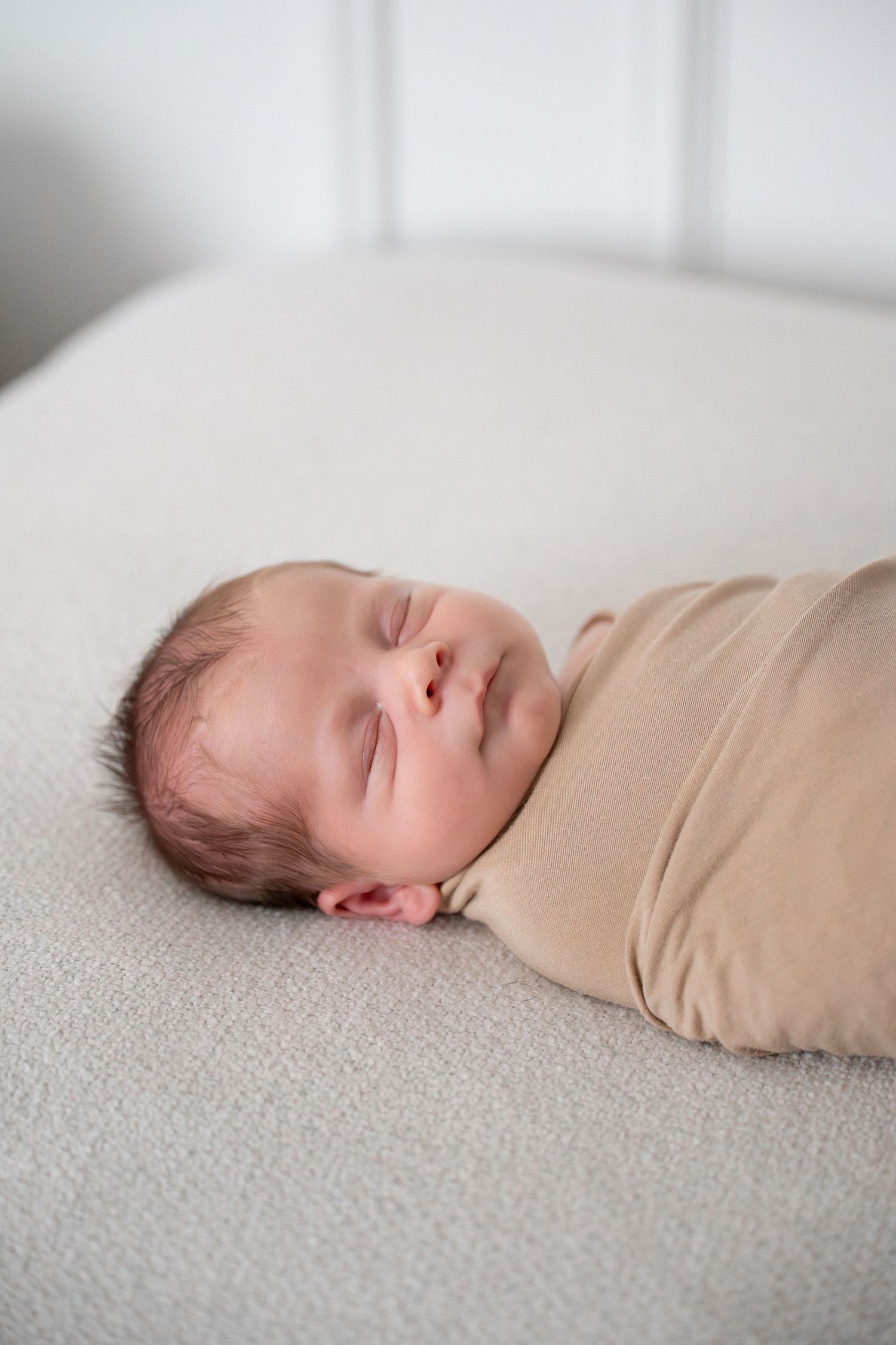 Sleeping newborn baby wrapped in tan swaddle resting peacefully on white textured surface.