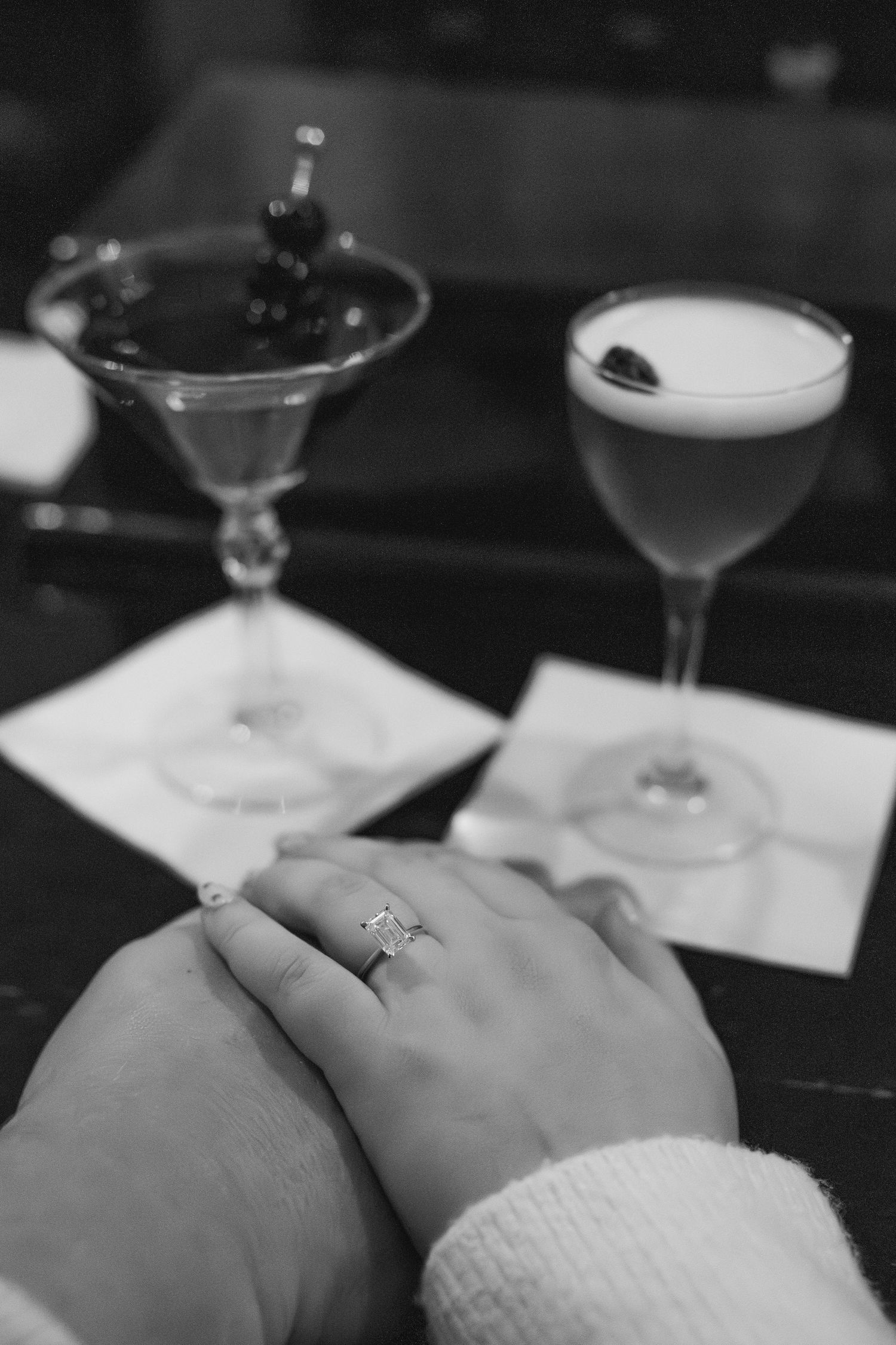 Close-up of hands with an engagement ring next to cocktail glasses on a dark table.