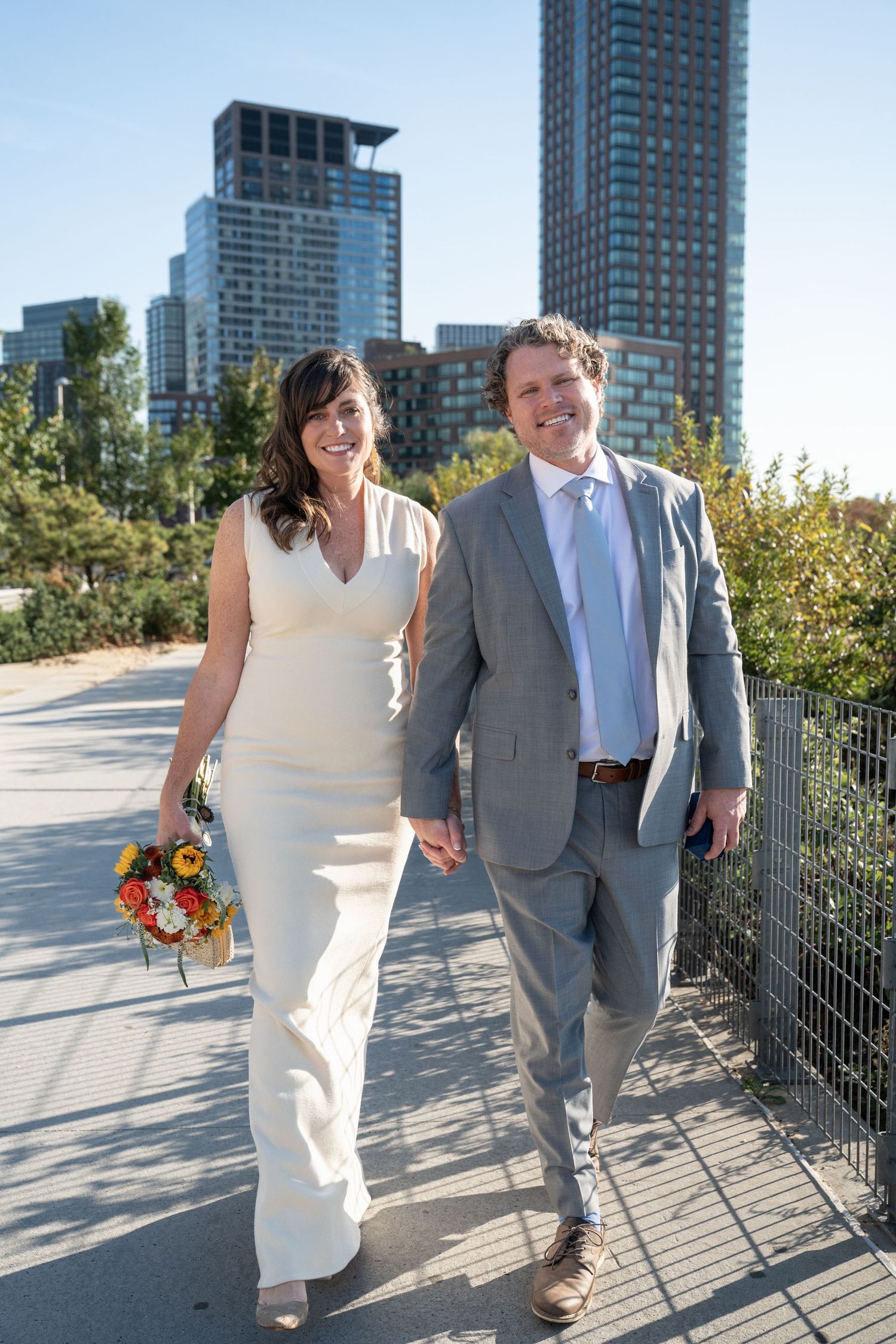 A couple walks together on a sunny day with city skyscrapers in the background.