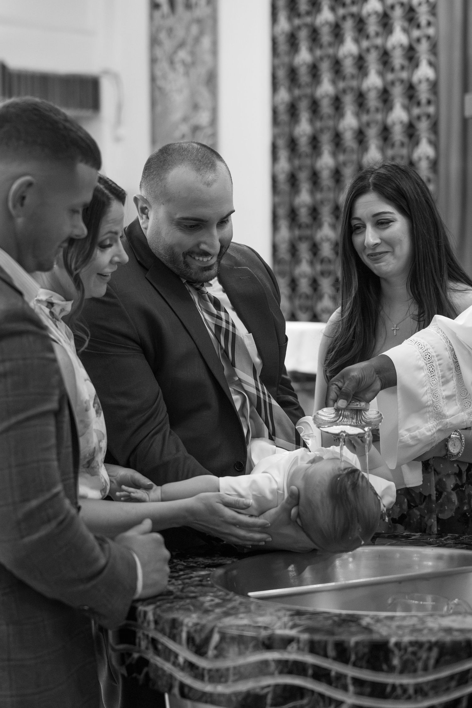 A black and white photo captures an intimate baptism moment as family members gather around a marble font.