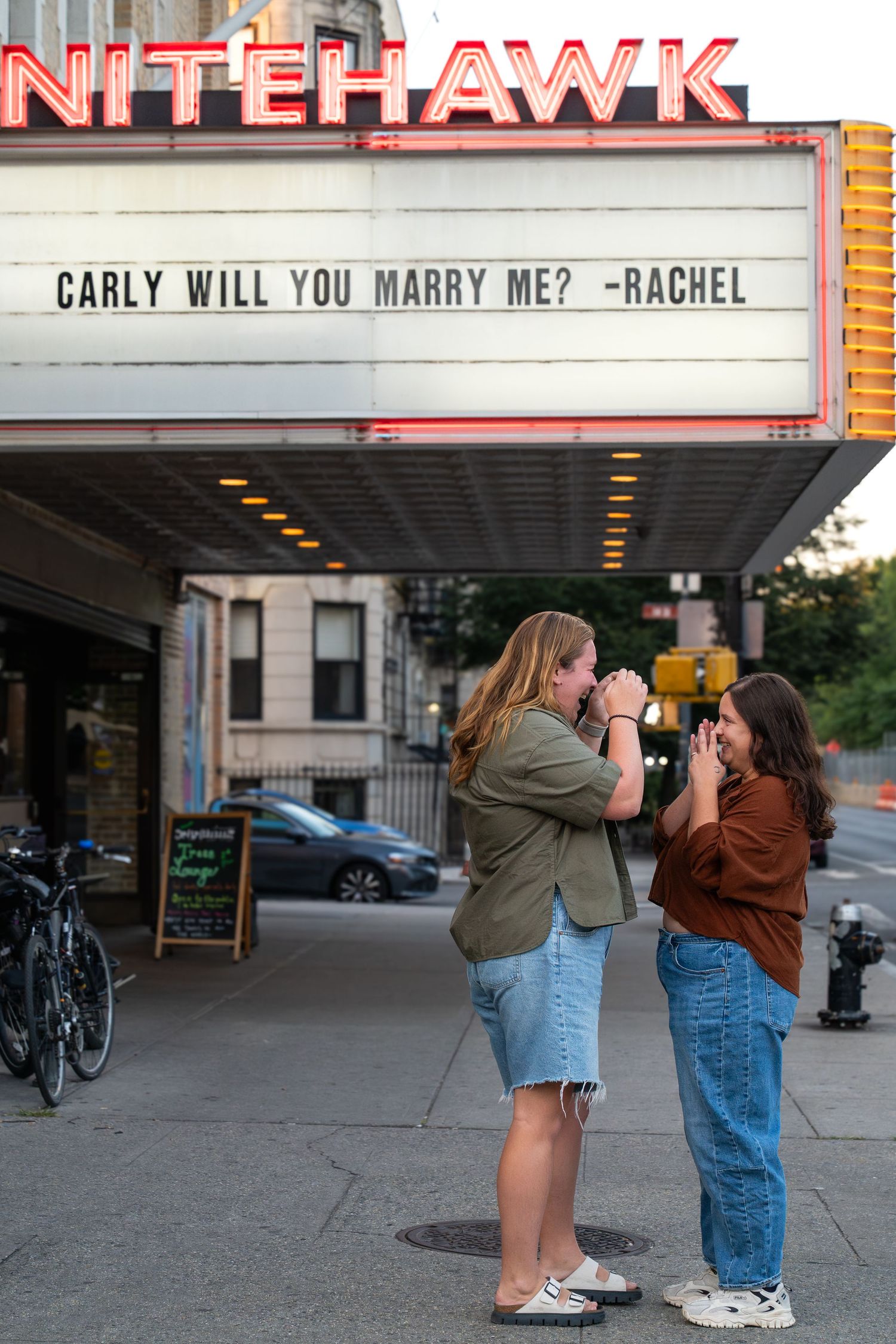 A theater marquee displays a marriage proposal message reading 'CARLY WILL YOU MARRY ME? -RACHEL' at the Nitehawk Cinema.