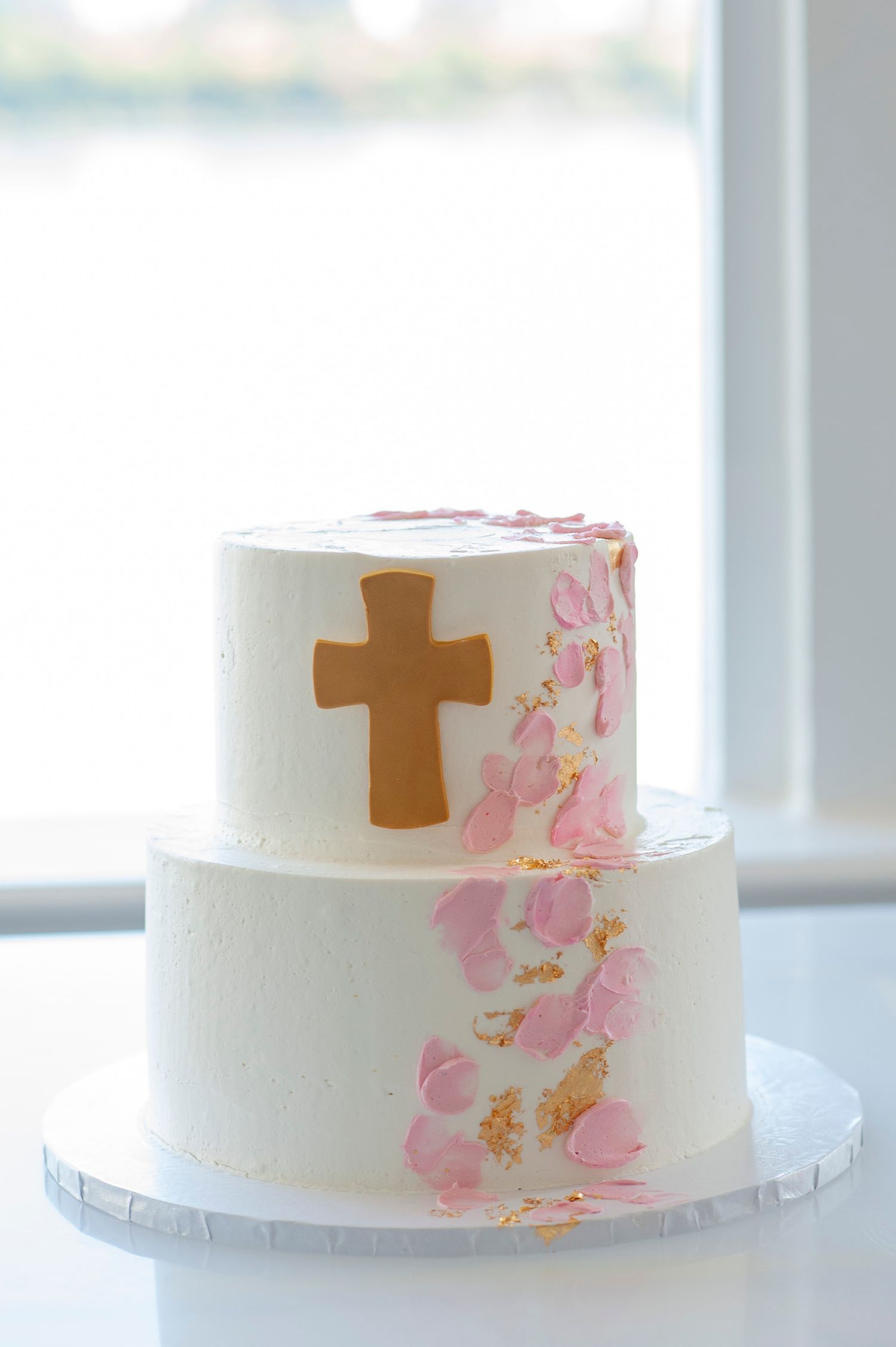White tiered cake with delicate pink floral designs and brown cross decorations for religious celebration.
