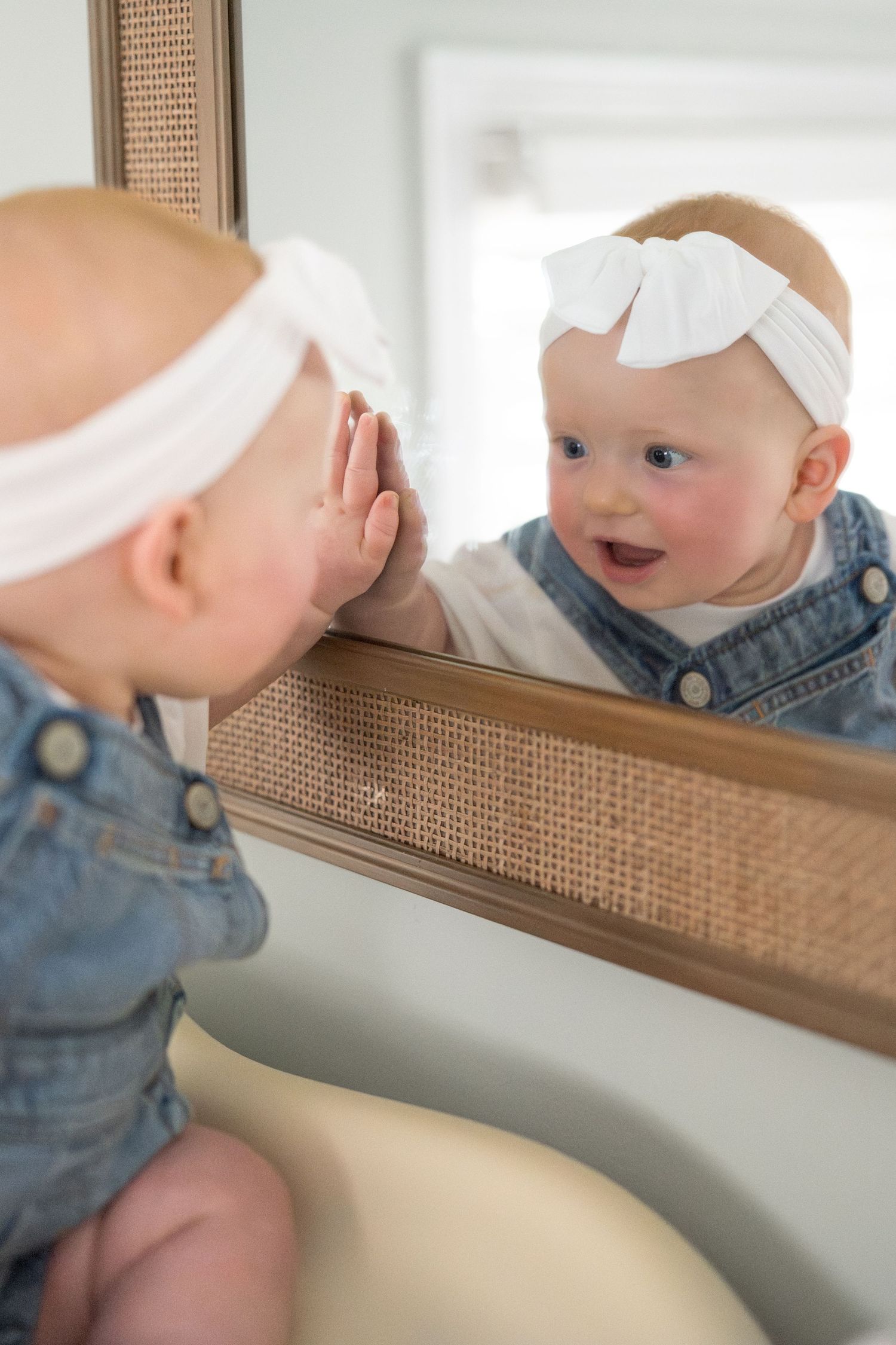 A small child in denim overalls explores their reflection in a bathroom mirror with curiosity.