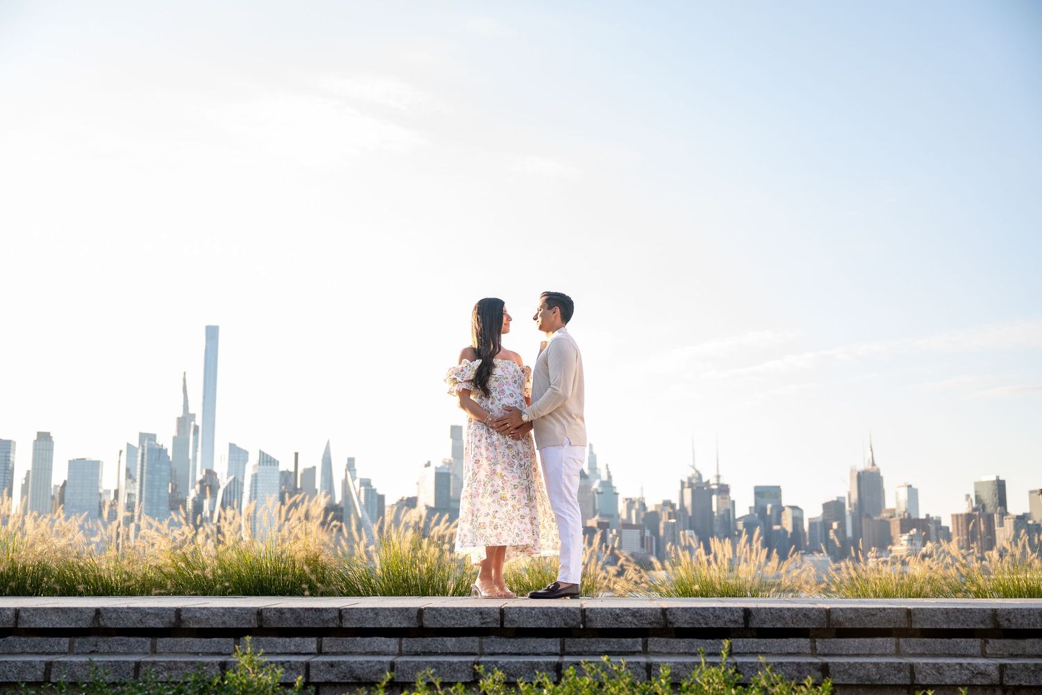 Couple embraces in front of New York City skyline with tall buildings and golden grass in foreground.