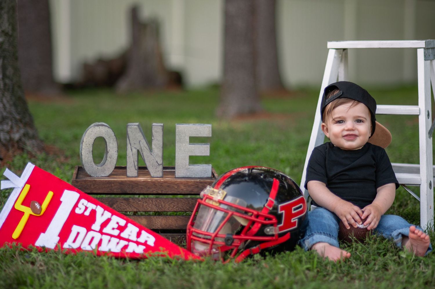 A small child sits by a ladder with decorative 'ONE' letters and red sports equipment on grass.