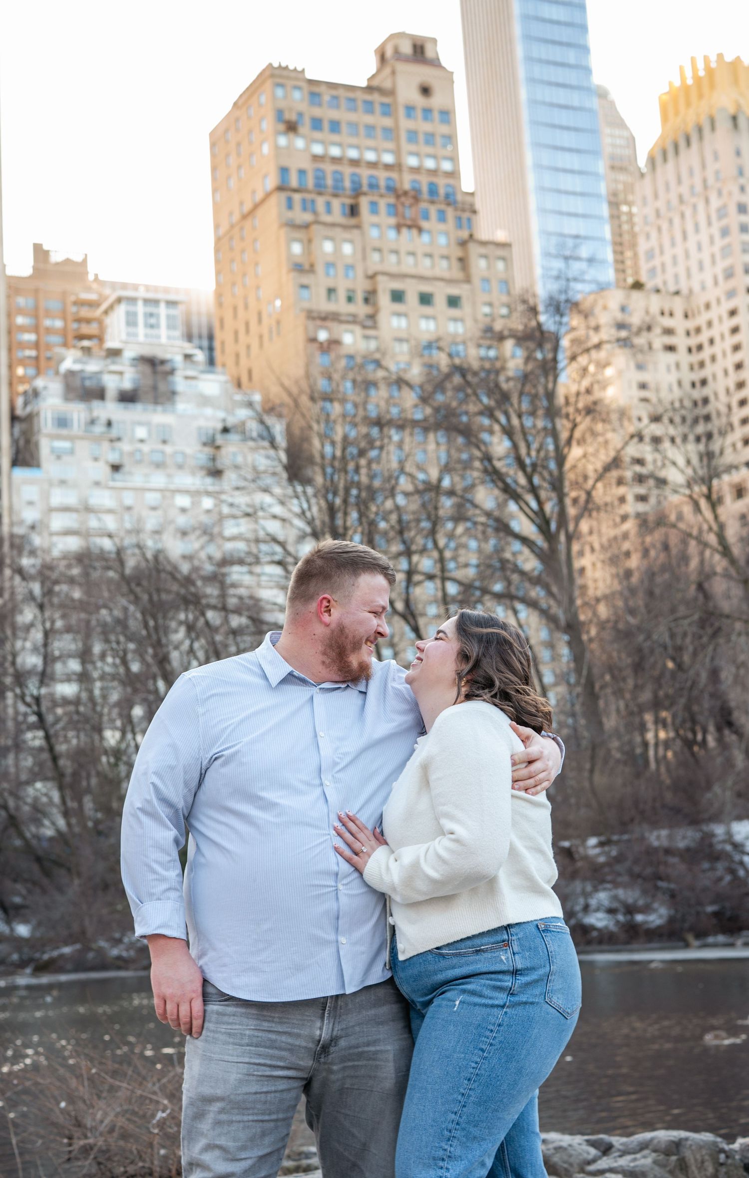 A romantic couple shares an intimate moment by the waterfront with city buildings in the background.