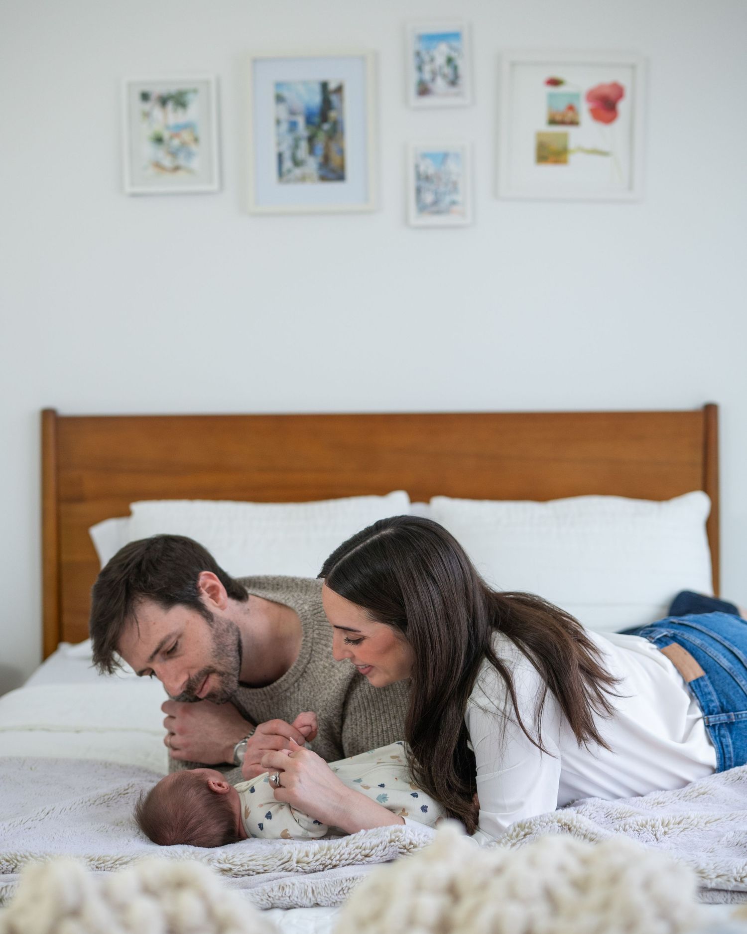 Family bonding with newborn baby on bed beneath framed artwork on white walls.