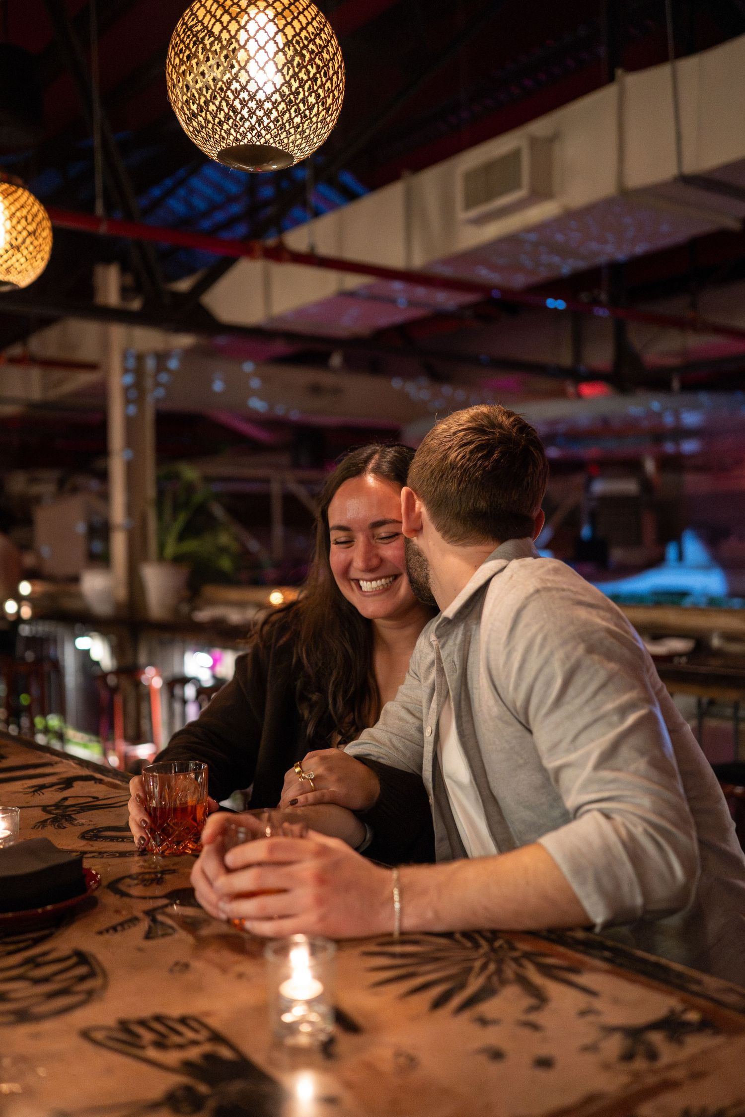 Two people share an intimate conversation at a dimly lit bar with decorative pendant lighting overhead.