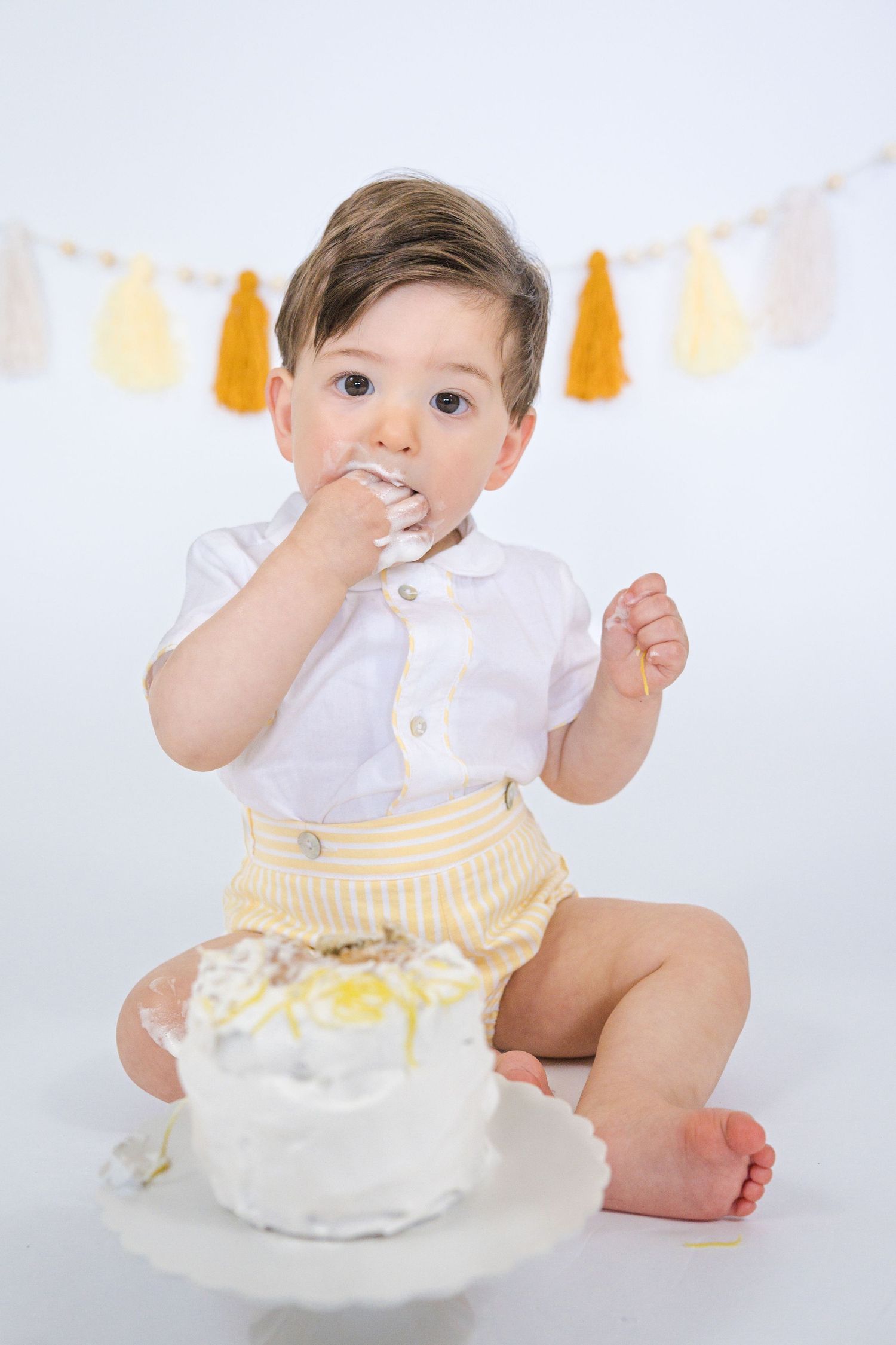 Baby in white and yellow outfit plays with birthday cake during cake smash photo session.