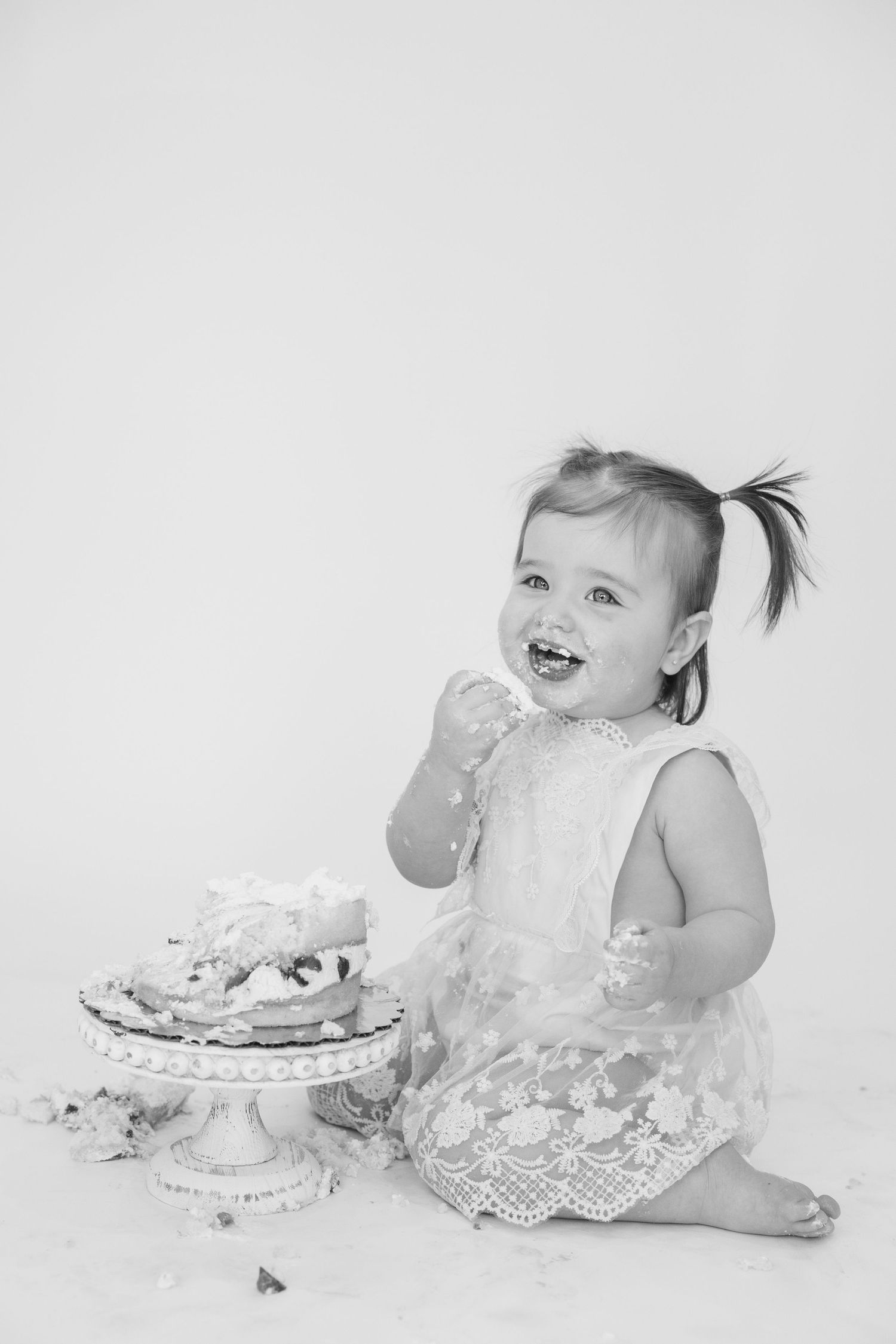 Baby in white dress laughing while playing with smashed cake during photo session.