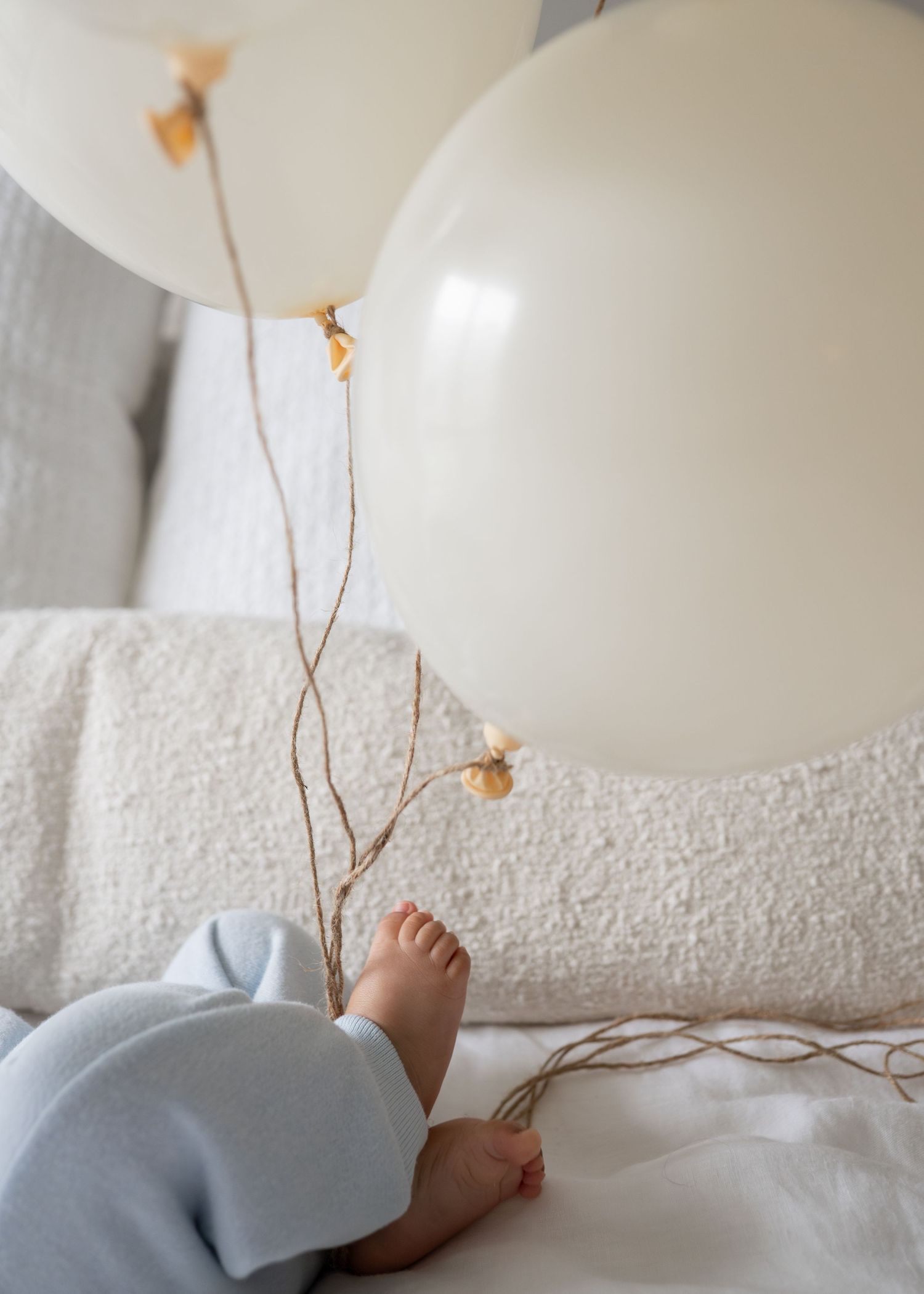 Baby feet in blue onesie next to cream balloons on white bed.