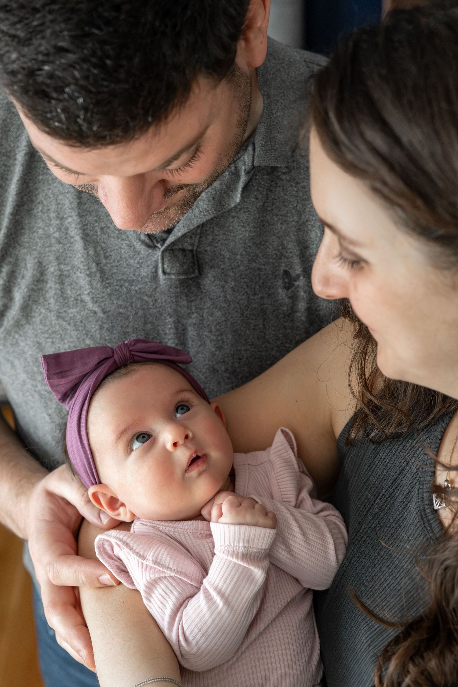 Parents sharing tender moments with their baby wearing pink clothing on gray couch.