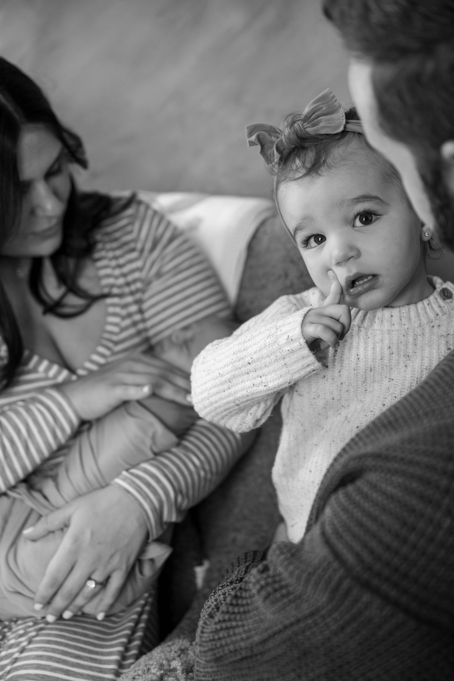 A black and white portrait of a baby in a knit sweater with a bow sitting with family members.