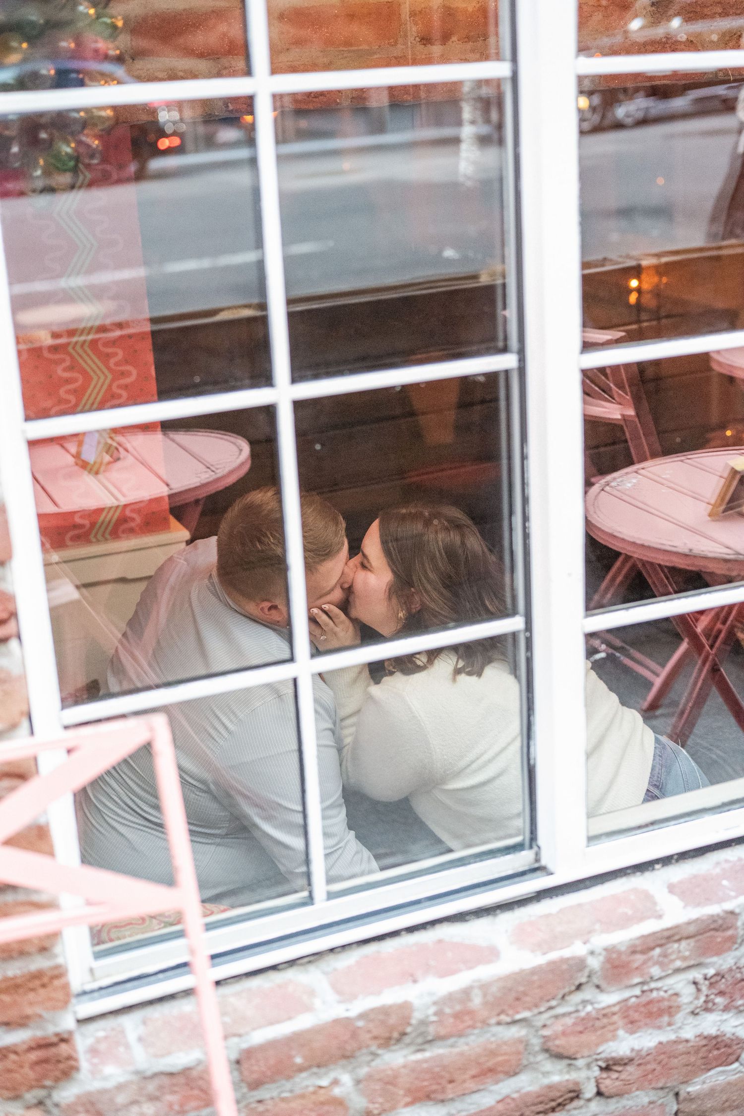 A couple shares a tender moment viewed through a window with reflections visible.