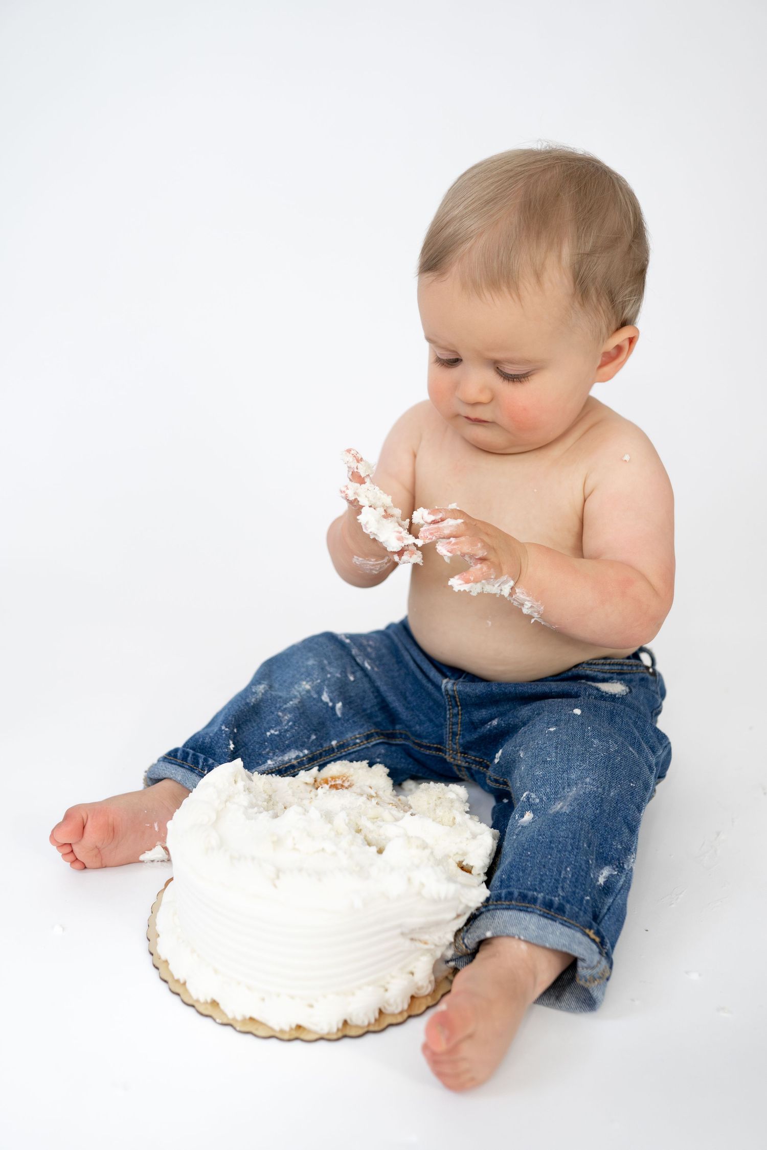 Baby in jeans smashing first birthday white cake, hands covered in frosting, sitting on white background.