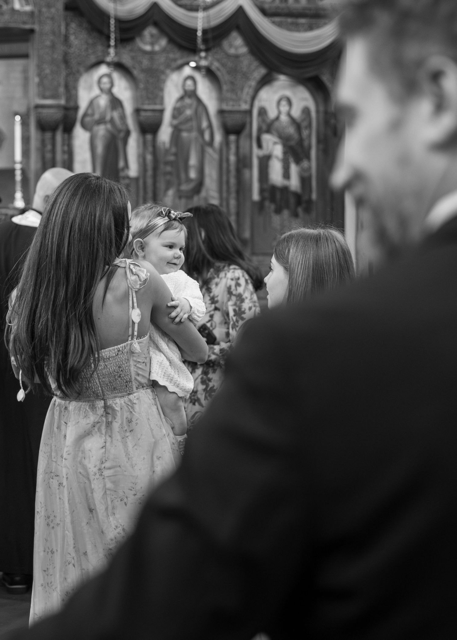 Black and white photo of a baby held by a woman in a church with religious icons in the background.