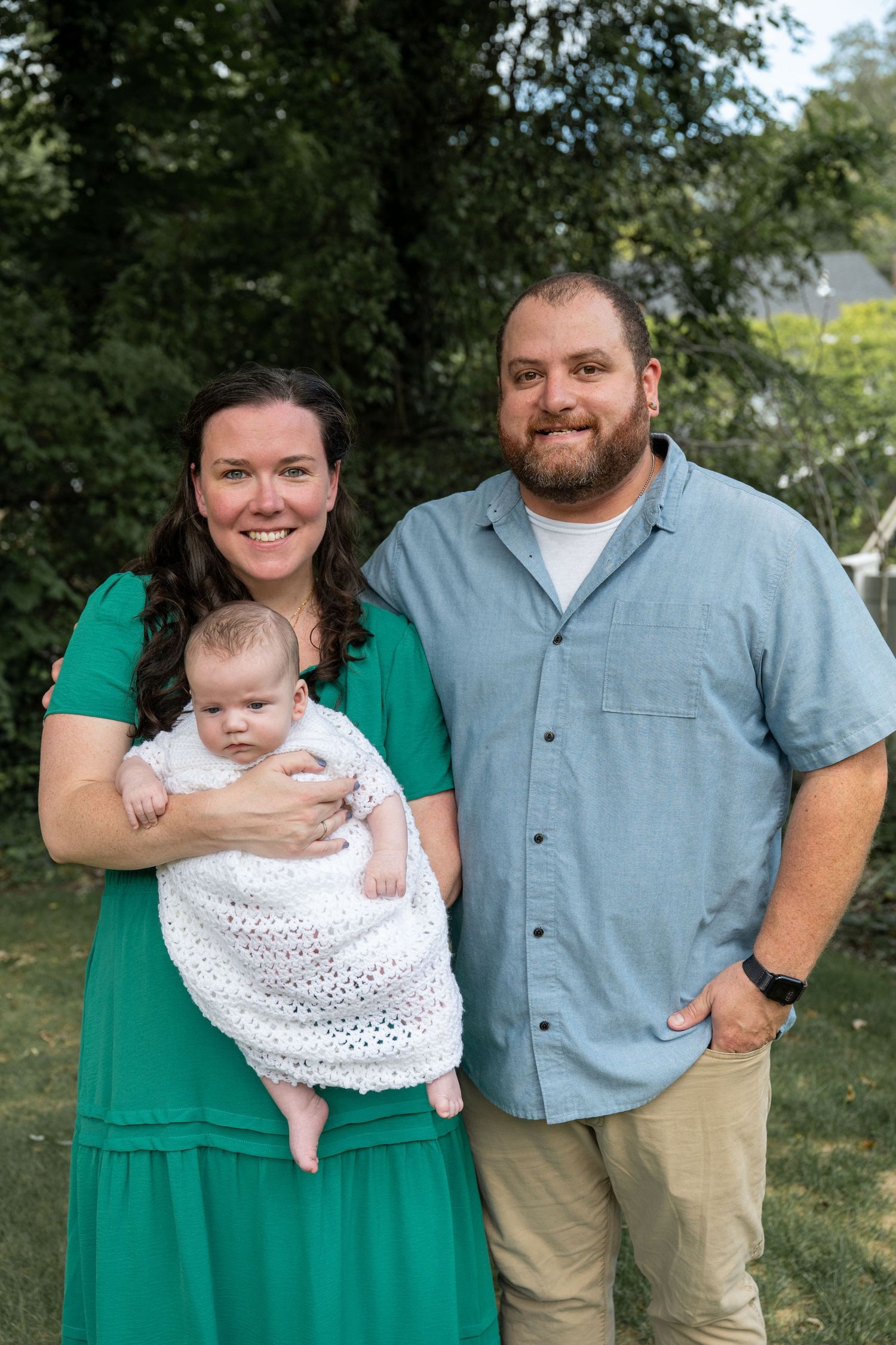 A family poses together outdoors with their baby wearing a white lace dress.